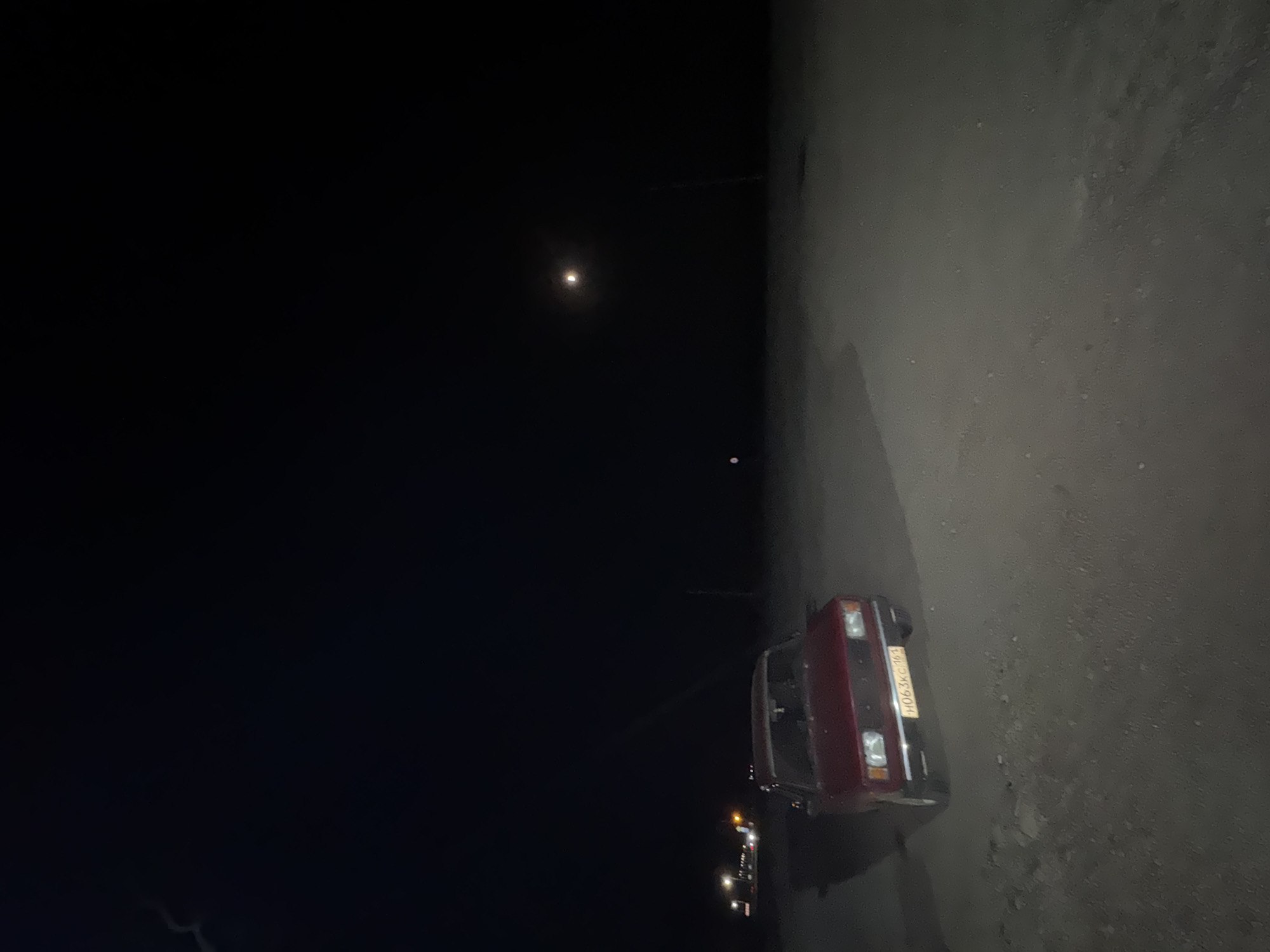 A dark, grainy, night-time photograph captures a red Lada car parked on a dirt road. The car's headlights are not illuminated. The license plate of the car reads "H063KC 161". In the distance, a faint light is visible, possibly a streetlamp or another vehicle's headlights. The sky is dark, with a faint, hazy moon or distant light source in the upper right quadrant. The overall setting appears to be a rural or undeveloped area, with the dirt road leading into darkness. The image does not provide enough information to determine the specific location within Yutin, Russia.
