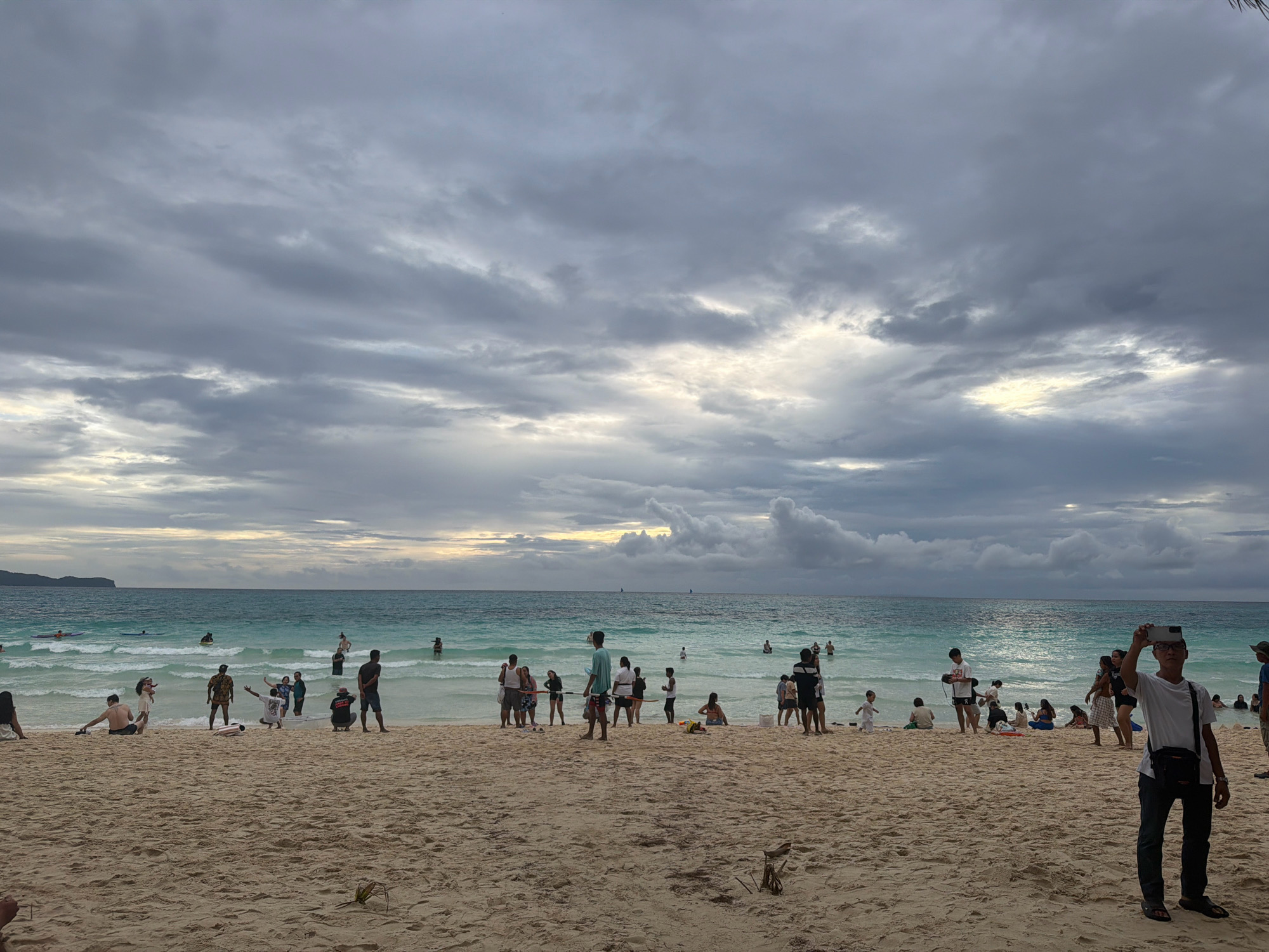 The image depicts a crowded beach scene in Malay, Philippines, likely on Boracay Island, given the characteristic turquoise waters and white sand. The sky is overcast with dramatic, dark grey clouds, suggesting a storm is approaching or has recently passed. Sunlight filters through a break in the clouds on the horizon, casting a soft glow on the ocean.
Numerous people are present on the sandy beach and in the shallow, clear turquoise water. On the sand, individuals are seen standing, sitting, and walking. Some are interacting with each other, while others appear to be relaxing or observing the surroundings. Several people are wading or swimming in the ocean, enjoying the water despite the cloudy weather.
In the foreground, on the right side of the frame, a man wearing a white t-shirt and dark pants is holding up a smartphone, seemingly taking a photograph or video. He has a dark crossbody bag slung over his shoulder and is wearing sandals.
Further into the scene, groups of people are scattered across the beach. Some are dressed in casual beach attire, including swimwear and t-shirts. Children are also visible, playing on the sand and in the water.
The ocean is a vibrant turquoise color, with gentle waves breaking near the shore. The water appears calm and inviting. In the distance, a faint outline of land can be seen on the left side of the horizon, possibly another island or a section of the coastline.
The beach itself is composed of light-colored, fine sand. Debris, such as small pieces of driftwood and dried leaves, are visible on the sand in the foreground. The overall impression is a lively beach environment with a significant number of visitors, under a moody, overcast sky.