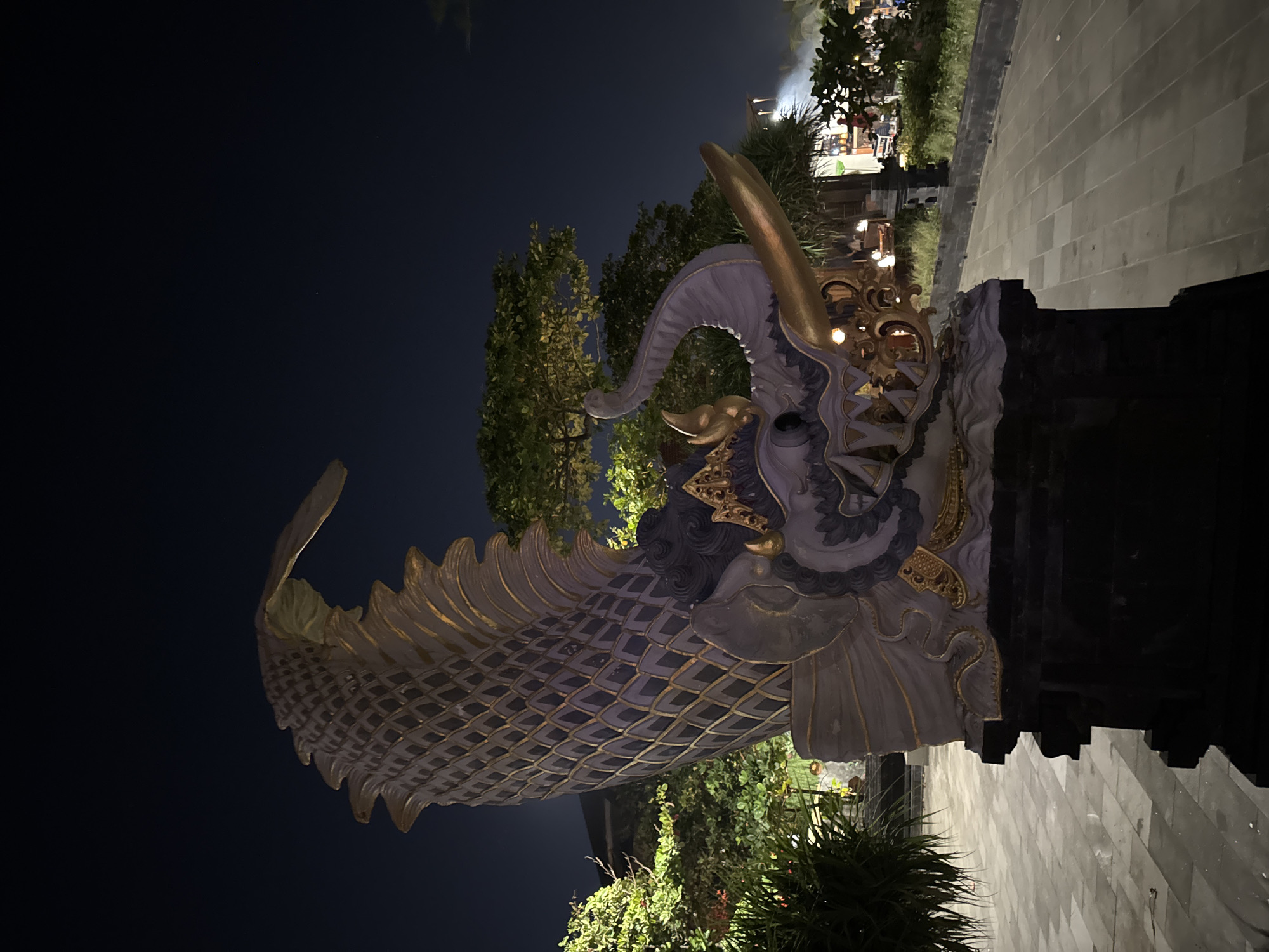 An imposing statue of a mythical creature, possibly a Naga or a dragon, stands prominently in an outdoor setting at night. The statue is elaborately decorated with scales and gold accents, featuring a long, serpentine body that curls upwards. The creature's head is detailed with fangs, a trunk-like appendage, and what appears to be a horn. The statue is illuminated by artificial lighting, casting shadows on the patterned stone ground. Lush green foliage surrounds the statue, with trees visible in the background. The night sky is dark, suggesting a late hour. The statue is a focal point in a public space, likely a tourist area in Kuta, Indonesia, as indicated by the architectural style and tropical vegetation. There are no people clearly visible in the immediate vicinity of the statue, but some indistinct figures and structures can be seen in the background, hinting at ongoing activity in the wider area. The overall scene conveys a sense of cultural art and nighttime ambiance.