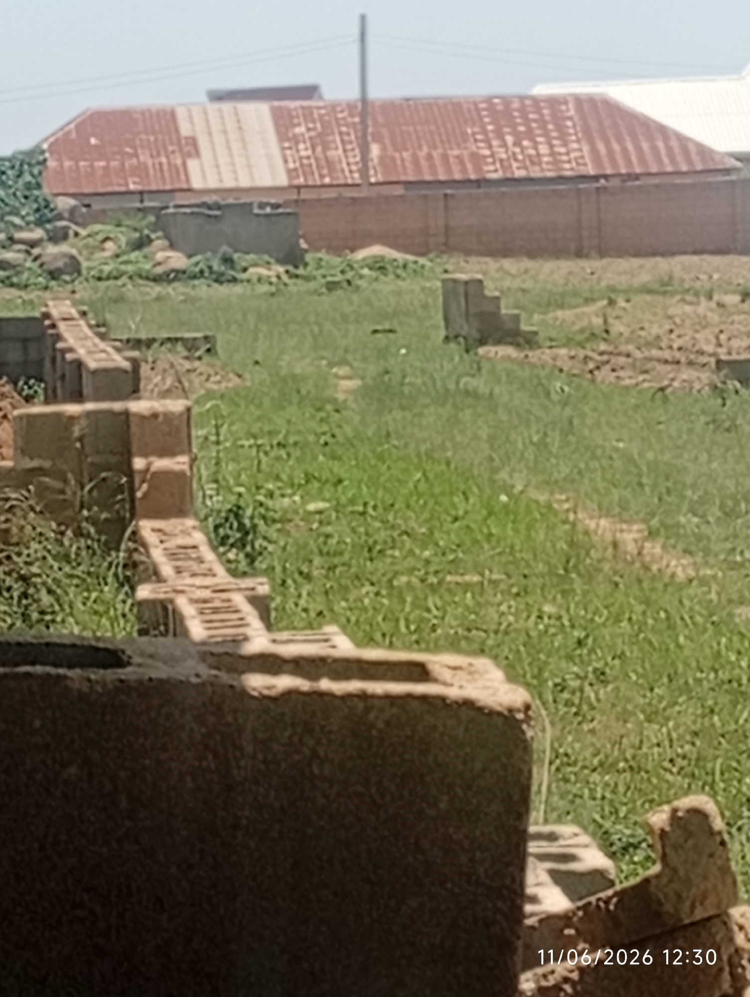 The image depicts an outdoor scene in DU, Nigeria, likely a construction site. In the foreground, partially visible brickwork suggests the beginnings of a structure, with a large, dark, textured object dominating the bottom left. Behind this, a row of partially constructed brick walls extends into the middle ground, with visible brick and mortar. Further back, a concrete step-like structure stands in a grassy area. The background features a long, brick wall, and beyond that, a building with a corrugated metal roof, showing signs of age and rust. The overall environment is characterized by dry grass, scattered debris, and a clear sky. The timestamp "11/06/2026 12:30" is visible in the bottom right corner. There are no people or animals visible in the image.