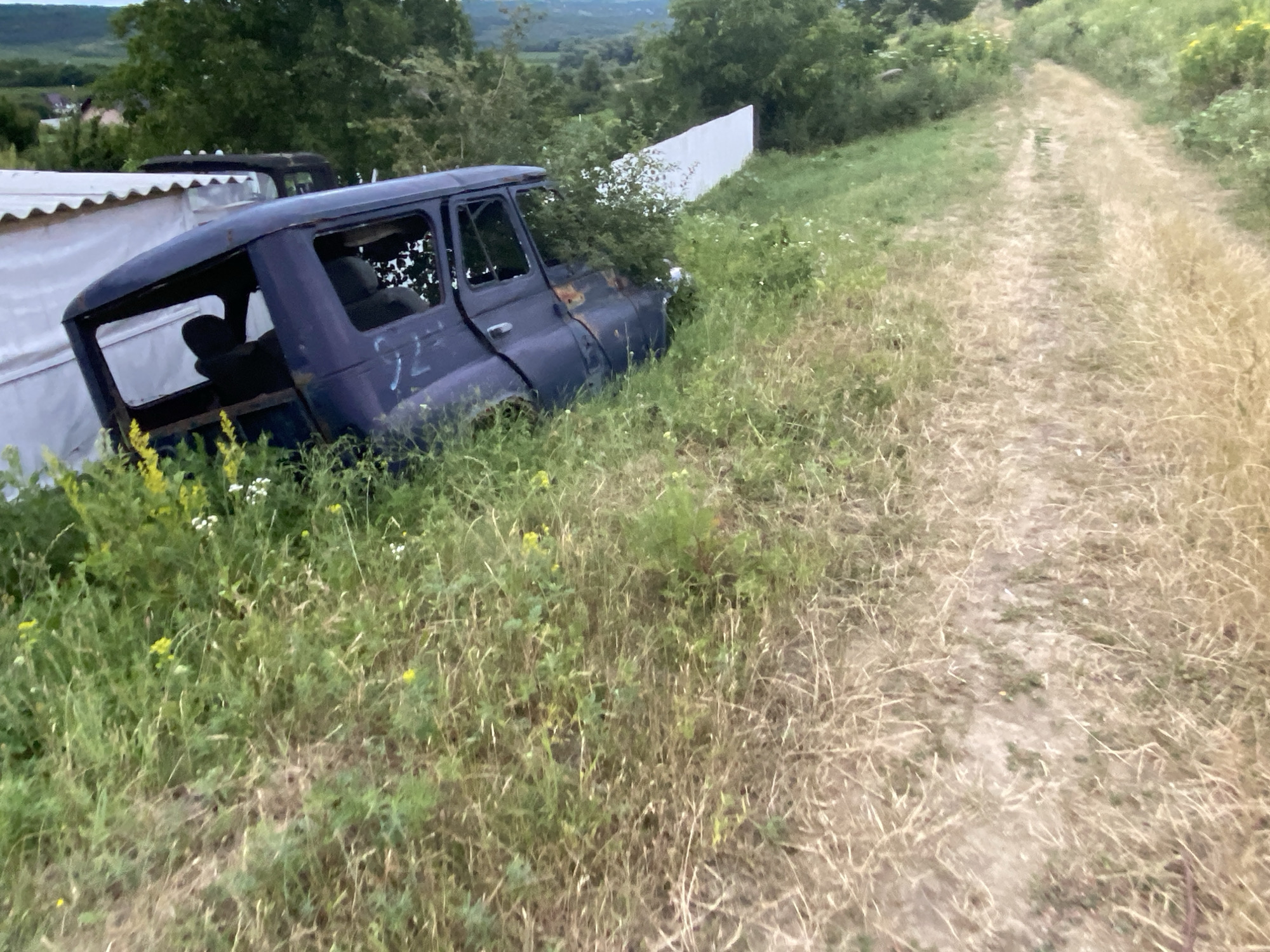 The image depicts an outdoor scene with an abandoned, dark blue van on the left, partially overgrown with grass and vegetation. The van appears to be in a state of disrepair, with its windows missing or broken. Behind the van, a white structure, possibly a temporary shelter or shed, is visible. To the right of the van, a dirt path or road winds through dry, tall grass and other greenery, leading into the background. The environment suggests a rural or semi-rural setting. The image was taken in or near Horodca, Moldova.