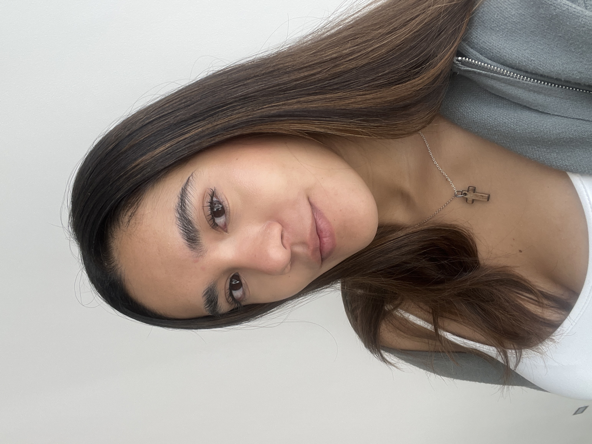 A woman is seen from the shoulders up, indoors against a light-colored wall. She has long, brown hair with highlights, and is wearing a white top and a gray zip-up hoodie. A silver necklace with a cross pendant is visible around her neck. Her expression is neutral. No location information is provided.
