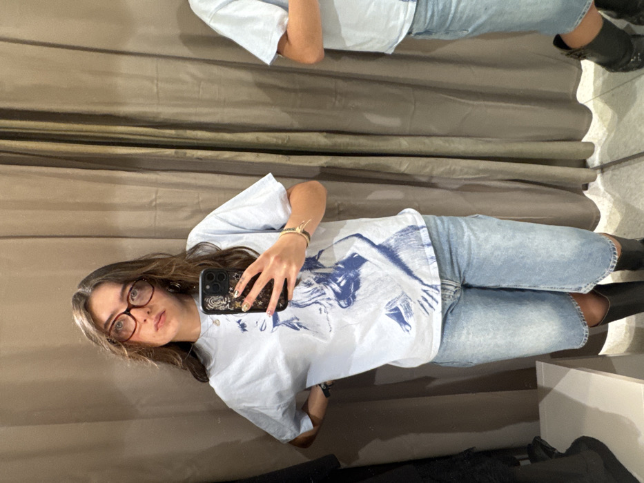A person with brown hair and glasses takes a selfie in front of a mirror. She wears a light blue t-shirt with a graphic design, light blue jeans, and black boots. Her phone has a decorated case. The mirror reflects another person wearing a similar outfit. The background consists of a neutral-colored curtain or wall, and a floor with a light-colored surface, possibly tile or linoleum.