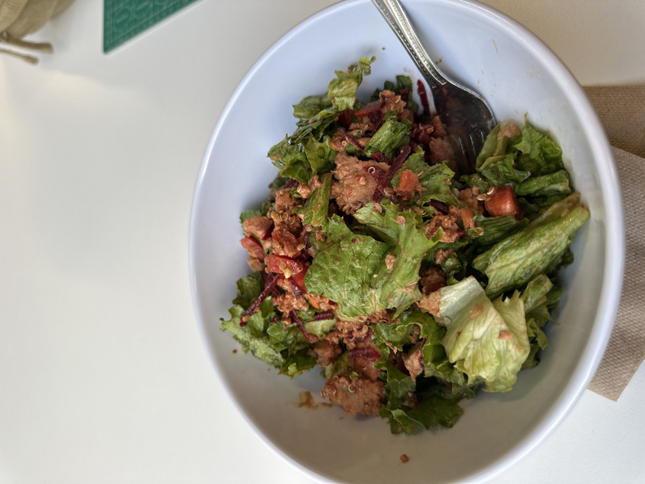 A vibrant salad fills a white bowl, accompanied by a silver fork. Green lettuce forms the base, topped with a mixture of ingredients, including diced tomatoes, shredded beets, and what appears to be quinoa or couscous. The salad is dressed, giving the leaves a slightly glossy appearance. The bowl sits on a white surface, next to a brown napkin or placemat.