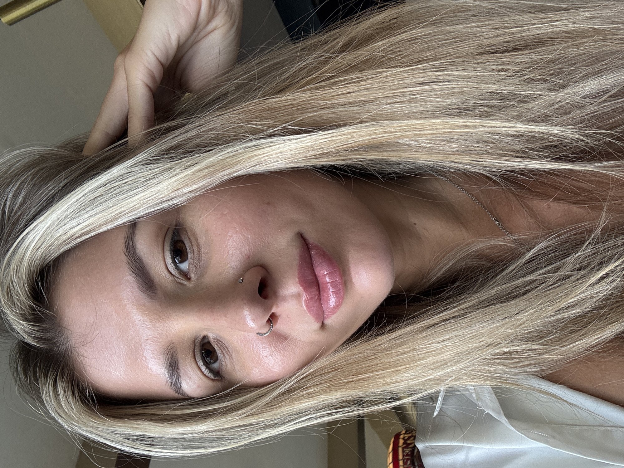 A close-up selfie shows the upper body of a woman with blonde hair. She is looking directly at the camera with her right hand on the side of her head, partially obscuring her hair. She is wearing a white shirt and a silver necklace. She has a nose ring on her left nostril. The background is blurred and appears to be an indoor setting.