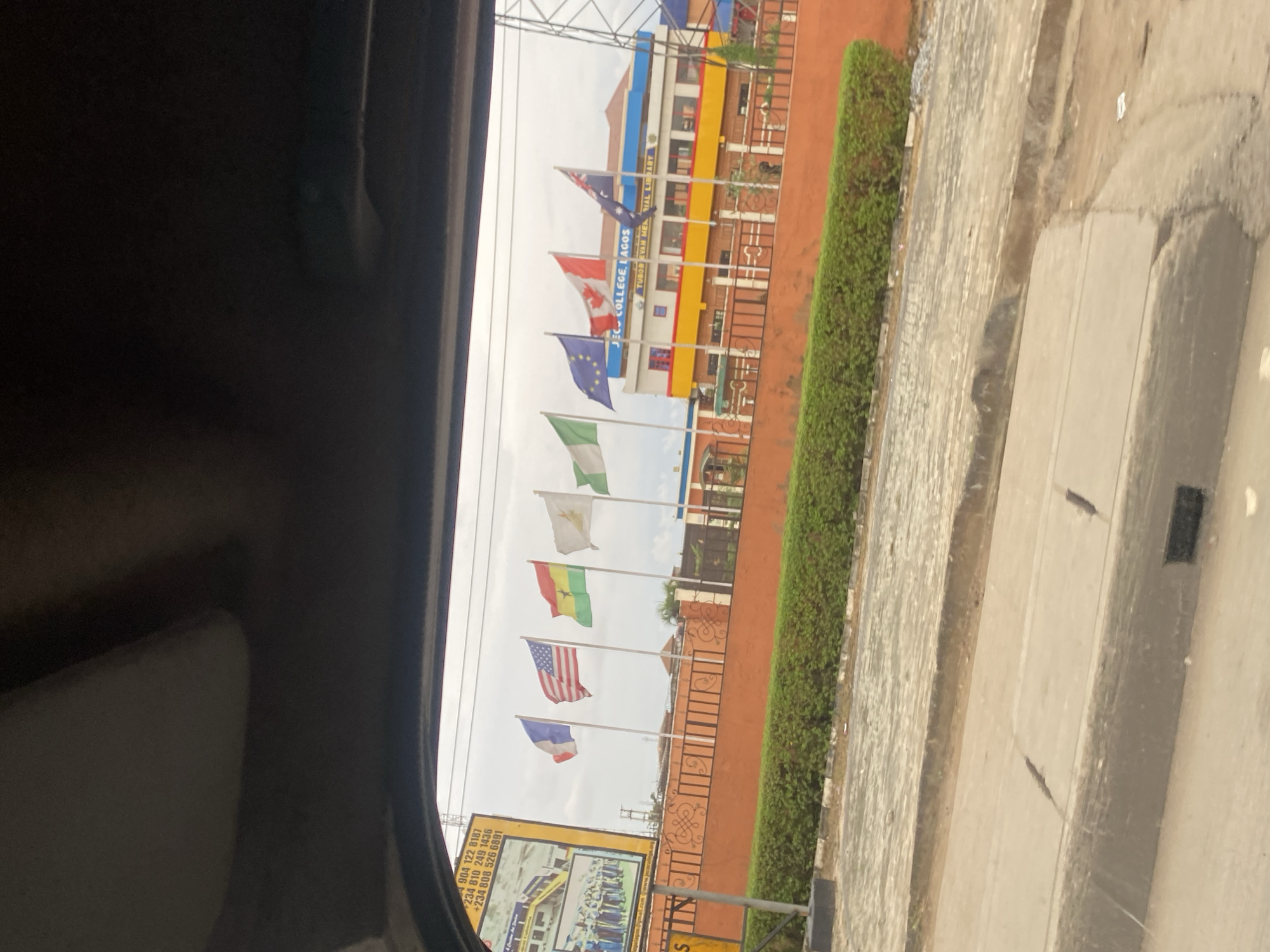 The image captures a view from inside a vehicle, looking out at the exterior of a school building identified as "JEC COLLEGE, LAGOS". The building has a distinctive yellow and blue facade with "TUNDE EVAN MEMORIAL LIBRARY" visible. In front of the school, several national flags are displayed on flagpoles: France, the United States, Ghana, a white flag with a symbol (possibly a crest or emblem), Nigeria, the European Union, Canada, and Australia. The flags are arranged in a line and are partially obscured by the vehicle's interior. A low, orange brick wall with decorative metalwork borders the school grounds, and a dense green hedge runs along the top of the wall. A paved area, possibly a walkway or courtyard, is visible behind the hedge. To the left of the scene, a large billboard with text and images of students is partially visible. The ground in the foreground appears to be a road or pavement with a drainage channel. The sky is overcast. The scene is likely a street in Lagos, Nigeria, based on the signage.