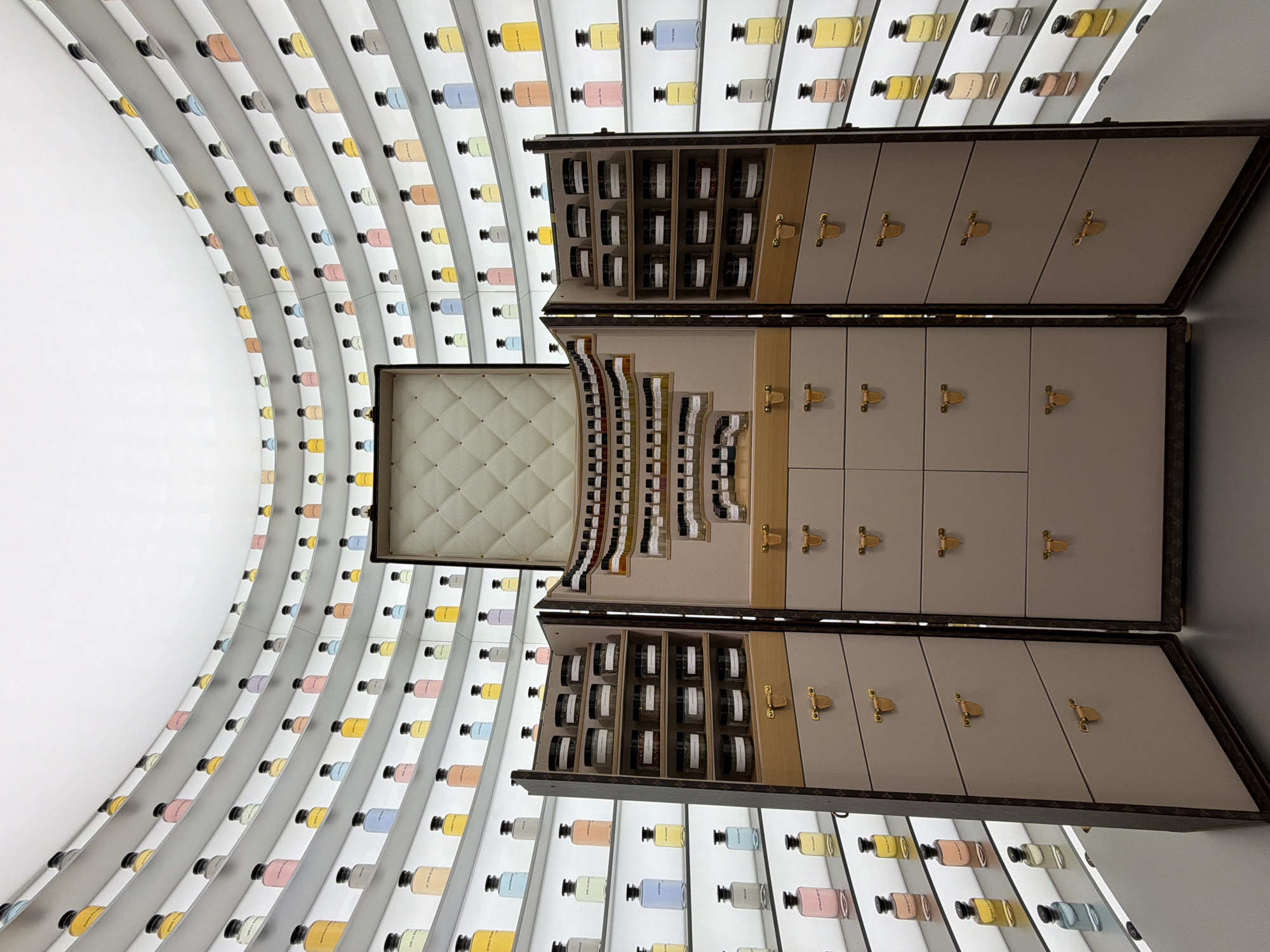 The image depicts an interior scene featuring a display cabinet and a wall lined with perfume bottles. The cabinet is in the foreground, with its doors open, revealing shelves filled with small jars. The doors are beige with brass-colored hardware. Behind the cabinet, a curved wall is lined with illuminated shelves, each holding a row of perfume bottles in various colors. The walls appear to be white. The location is Jing'an District, China.