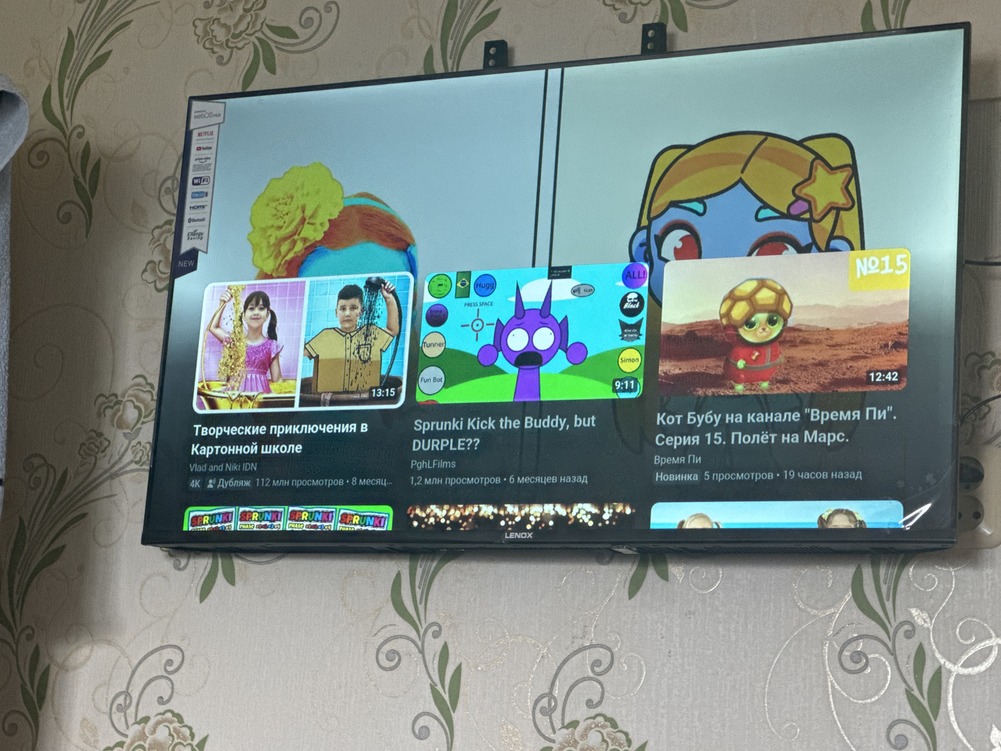 The image shows a television screen displaying various YouTube videos. The TV is mounted on a wall with floral wallpaper. On the left side of the screen, a video titled "Творческие приключения в Картонной школе" (Creative Adventures in Cardboard School) is featured, with a thumbnail showing two children. Below this, the video has 4K resolution and 112 million views, posted 8 months ago by "Vlad and Niki IDN".
In the center of the screen, a video titled "Sprunki Kick the Buddy, but DURPLE??" is displayed. Its thumbnail shows a purple cartoon character with big eyes. This video has 1.2 million views and was posted 6 months ago by "PghlFilms".
On the right side, a video titled "Кот Бубу на канале 'Время Пи'. Серия 15. Полёт на Марс." (Cat Booboo on the "Time Pi" channel. Episode 15. Flight to Mars.) is shown. The thumbnail features a cartoon turtle character. This video is a new release with 5 views and was posted 19 hours ago.
A sidebar on the far left of the TV displays app icons including Netflix, YouTube, Prime Video, and Wi-Fi. The television is a smart TV with "webOS Hub" indicated at the top. The bottom of the television has a "LENOX" logo. The overall scene suggests a living room environment.
