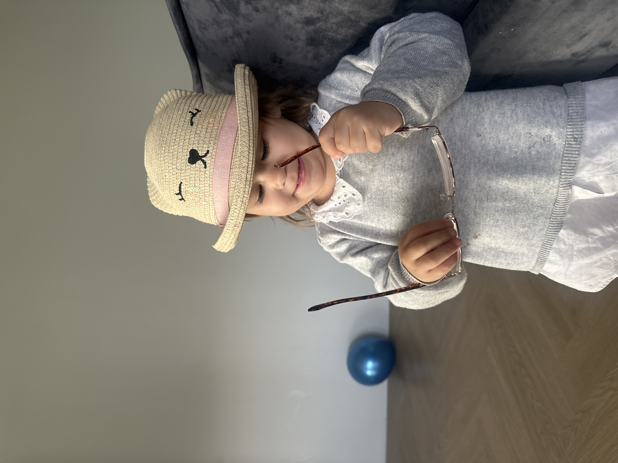 An indoor, top-down shot taken in Kharnavy, Russia, features a young child, likely a girl, playfully interacting with a pair of eyeglasses while lying on a dark gray sofa or cushion. The child wears a light-colored straw hat with a black bunny face design and a pink ribbon band. She is dressed in a light gray long-sleeved sweater with a white Peter Pan-style collar, over a white dress or shirt.
The child holds the eyeglasses with both hands, one arm of the glasses playfully near her mouth or nose, and exhibits a sweet, curious smile, conveying a sense of happiness and engagement.
The background shows a plain light gray wall and light brown wooden flooring. A single blue balloon is visible on the floor in the lower right corner of the frame. The image's orientation is such that the child appears horizontal across the frame. No text is visible in the image.