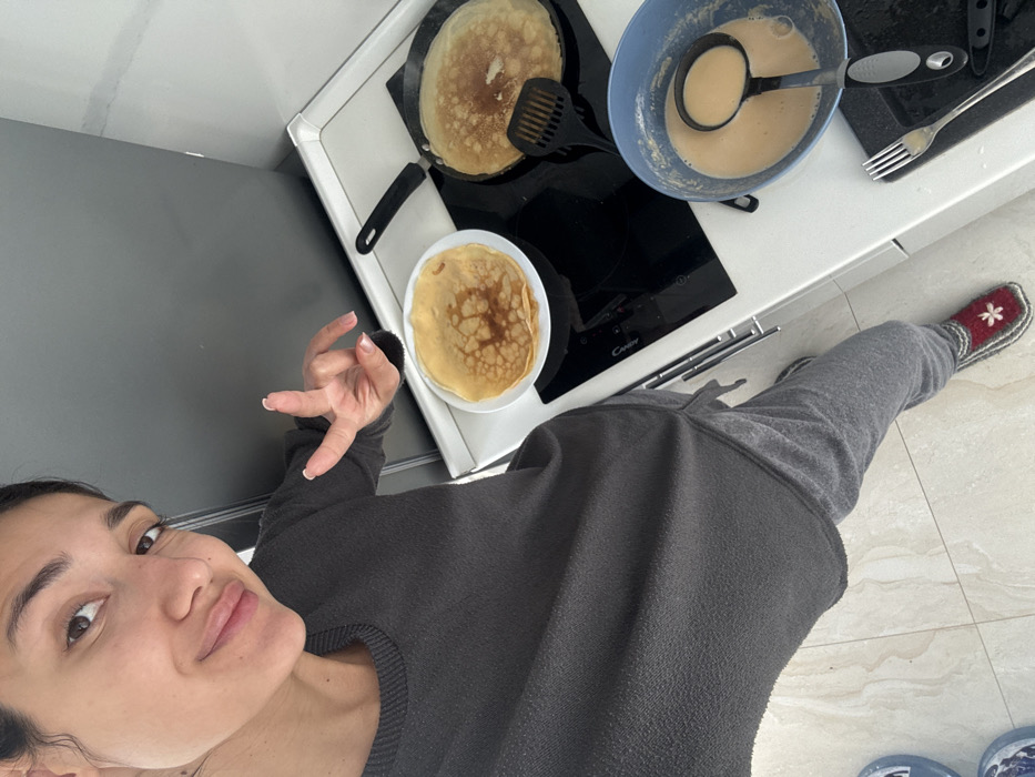 This image depicts an indoor kitchen environment located in Budva, Montenegro. An adult female is the primary subject, occupying the foreground and left side of the frame. She has dark hair and eyes, and is wearing a dark grey long-sleeved top and grey sweatpants. Her visible left foot is clad in a red and white knitted slipper. She is smiling directly at the camera, and her right hand is raised, forming a two-fingered "peace" gesture.
The background reveals a modern kitchen setup. A white tiled floor is visible in the foreground. A white countertop extends across the mid-ground, featuring a black induction or ceramic stovetop, identified by the "Candy" brand label. On the left burner, a cooked golden-brown crepe or pancake rests on a white round plate. On the right burner, a second crepe is actively cooking in a black frying pan, with a black slotted spatula positioned within it. To the right of the stovetop, a light blue mixing bowl contains liquid batter and a black ladle. A silver fork and a black utility knife are placed on the countertop near the bowl. A dark grey refrigerator is visible in the upper background.