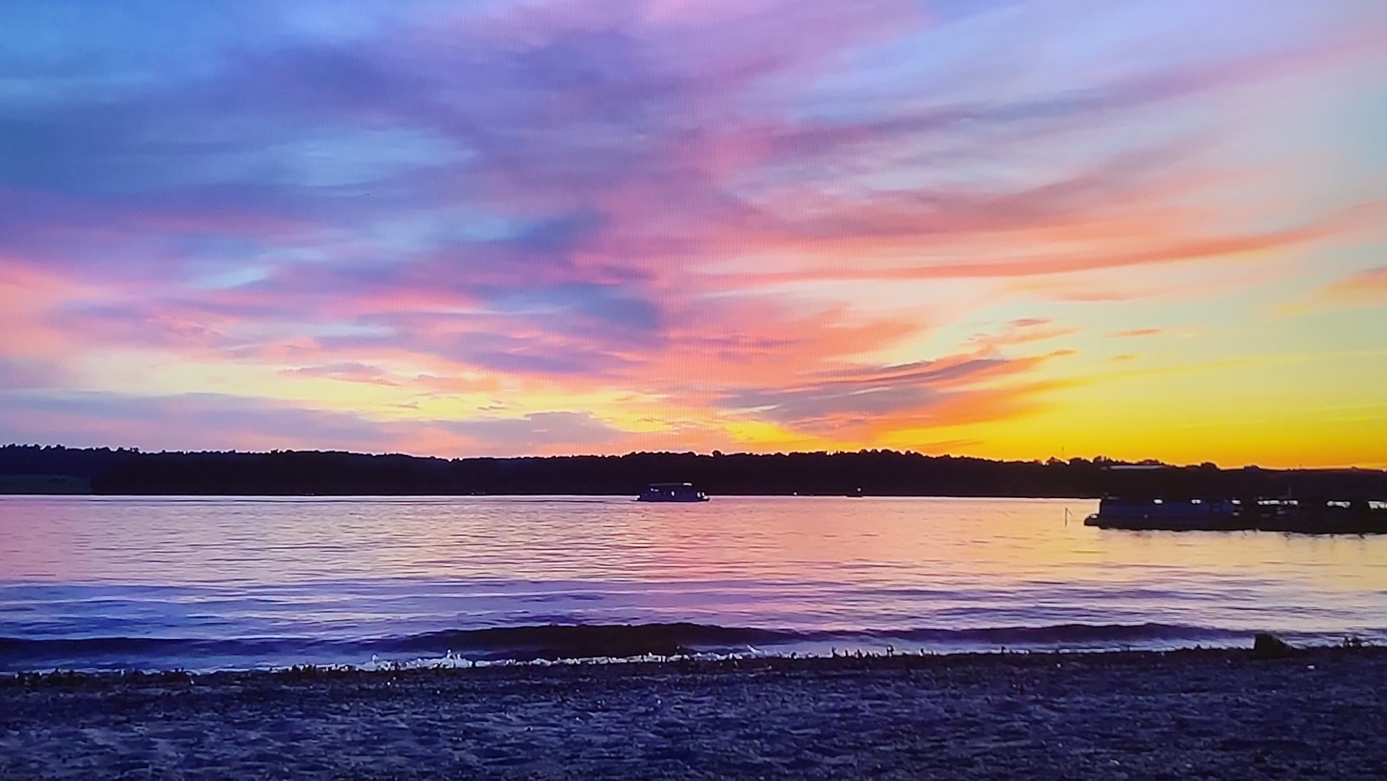 The image shows a sunset over a body of water. The sky is filled with colorful clouds in hues of pink, purple, orange, and yellow. A line of dark trees sits on the horizon, separating the sky from the water. In the water, there appears to be a small boat or structure. Closer to the viewer, there is a beach with sand.
It is impossible that the picture was taken in Yerevan, Armenia because Yerevan is a landlocked city without a coastline.