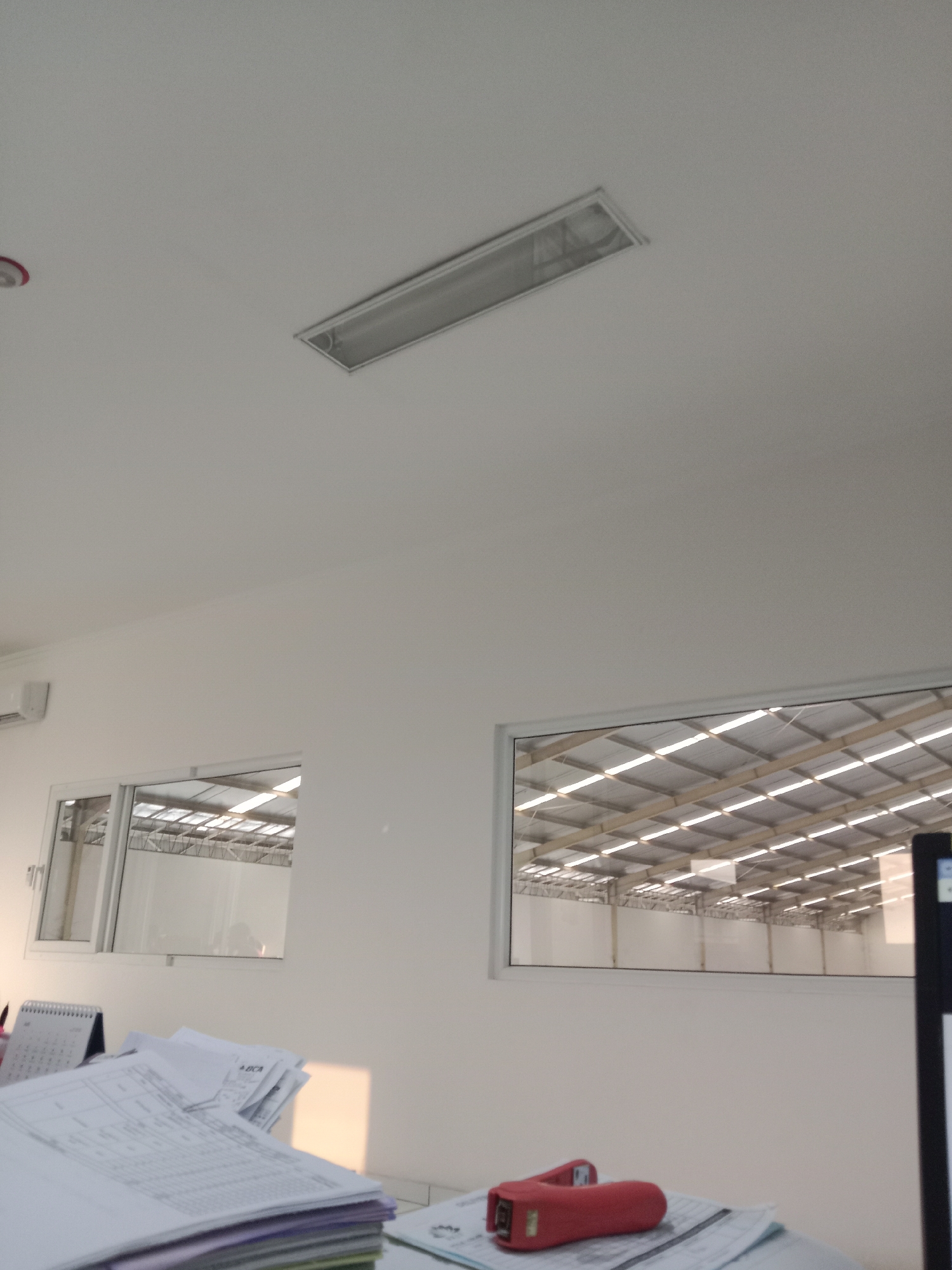 This image depicts the interior of a modern office space, likely a warehouse or industrial facility, with a focus on the ceiling and windows. The ceiling is a plain white surface with a rectangular fluorescent light fixture installed centrally. Below the ceiling, there are two windows. The left window is smaller and shows a reflection of the interior, including what appears to be an air conditioning unit on the wall. The larger right window offers a view into a spacious, brightly lit industrial hall with a high ceiling supported by a grid of wooden beams and corrugated metal roofing. Sunlight streams through the translucent roofing panels, creating a pattern of light and shadow. In the foreground, a desk is visible with stacks of papers and a red handheld scanner. The overall impression is one of a clean, functional workspace. No specific landmarks or details are provided to pinpoint the exact location in Semarang, Indonesia, beyond it being an interior office or industrial setting.