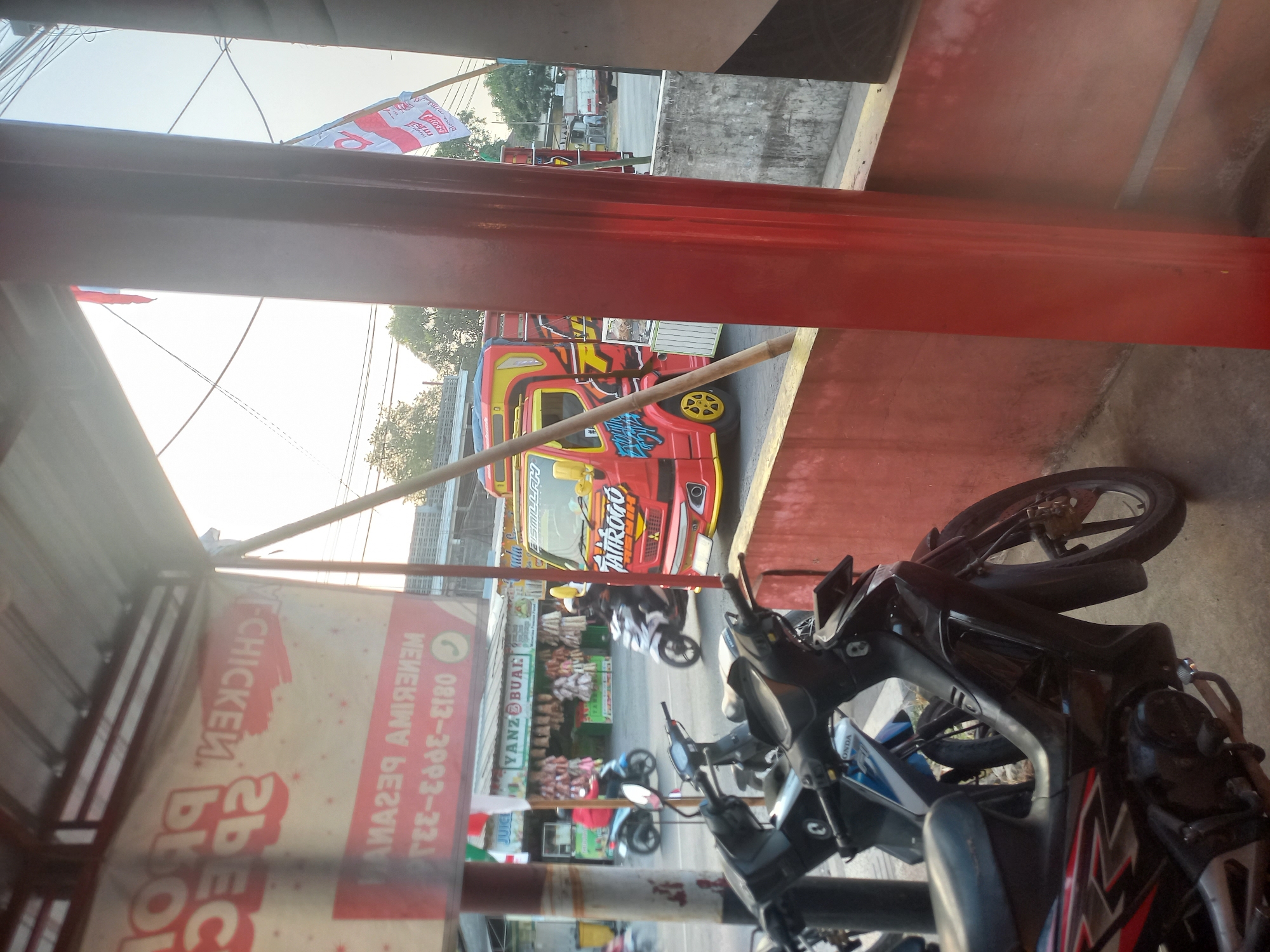 The image depicts a street scene in Bedah Lawak, Indonesia. In the foreground, several parked motorcycles are visible, one black and red motorcycle in the lower left quadrant, and another partially visible blue and white motorcycle behind it. A vibrant red and yellow truck with "Tattrogo" written on its side is a prominent feature in the midground, positioned on the street. The truck has colorful graphics and appears to be driving or parked. Behind the truck, a small market stall with various items hanging can be seen, identified by a sign that reads "YANZ BUAH". A banner with red lettering, partially obscured and inverted, is also visible on the left side of the image, with legible text including "CHICKEN" and a phone number "0813-333-333". In the background, several utility poles and wires crisscross the sky, and trees are visible. The overall lighting suggests it is daytime, possibly late afternoon given the warm glow. The scene appears to be a typical street environment in a bustling Indonesian town, with commercial activity and traffic.