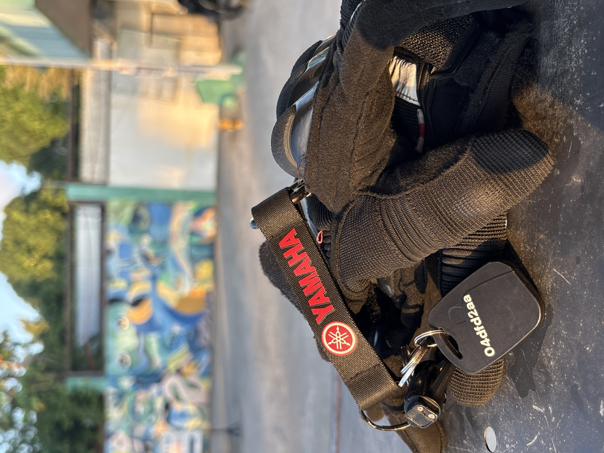 A close-up shot captures a pair of black motorcycle gloves, a Yamaha-branded strap, and a key fob lying on a dark, textured surface, possibly a table or pavement. The Yamaha strap features the brand's logo and name in red. A black key fob with the alphanumeric code "O4dfd2aa" is attached to the strap with a keyring. In the background, a vibrant mural adorns a wall, with lush green trees and a building visible under a bright sky. The scene is lit by warm, golden sunlight, suggesting it's either sunrise or sunset. The image appears to be taken in Tagum City, Philippines, as indicated by the presence of a local key fob design often seen in the area. The focus is sharp on the foreground objects, while the background is softly blurred, emphasizing the motorcycle gear.