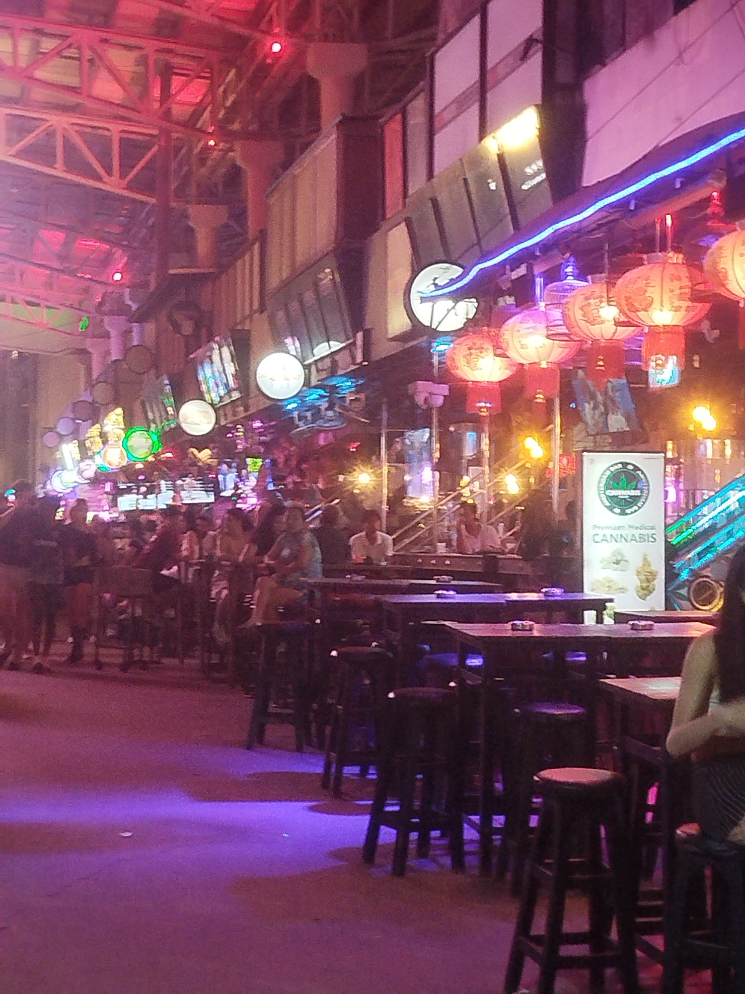 The image depicts a lively street scene at night, likely in an entertainment district, with a prominent focus on a bar or restaurant area. The setting is illuminated by a vibrant mix of neon lights, red and pink ambient lighting, and traditional red lanterns hanging overhead. A row of tables with stools is visible, suggesting an outdoor seating arrangement. Several people are seated at the tables, engaged in conversation and appearing relaxed. In the background, a sign prominently displays "Premium Medical CANNABIS," indicating the nature of some establishments in the area. The architecture suggests a multi-story building with industrial elements, possibly a converted warehouse or an open-air market. The overall atmosphere is one of a bustling nightlife scene.