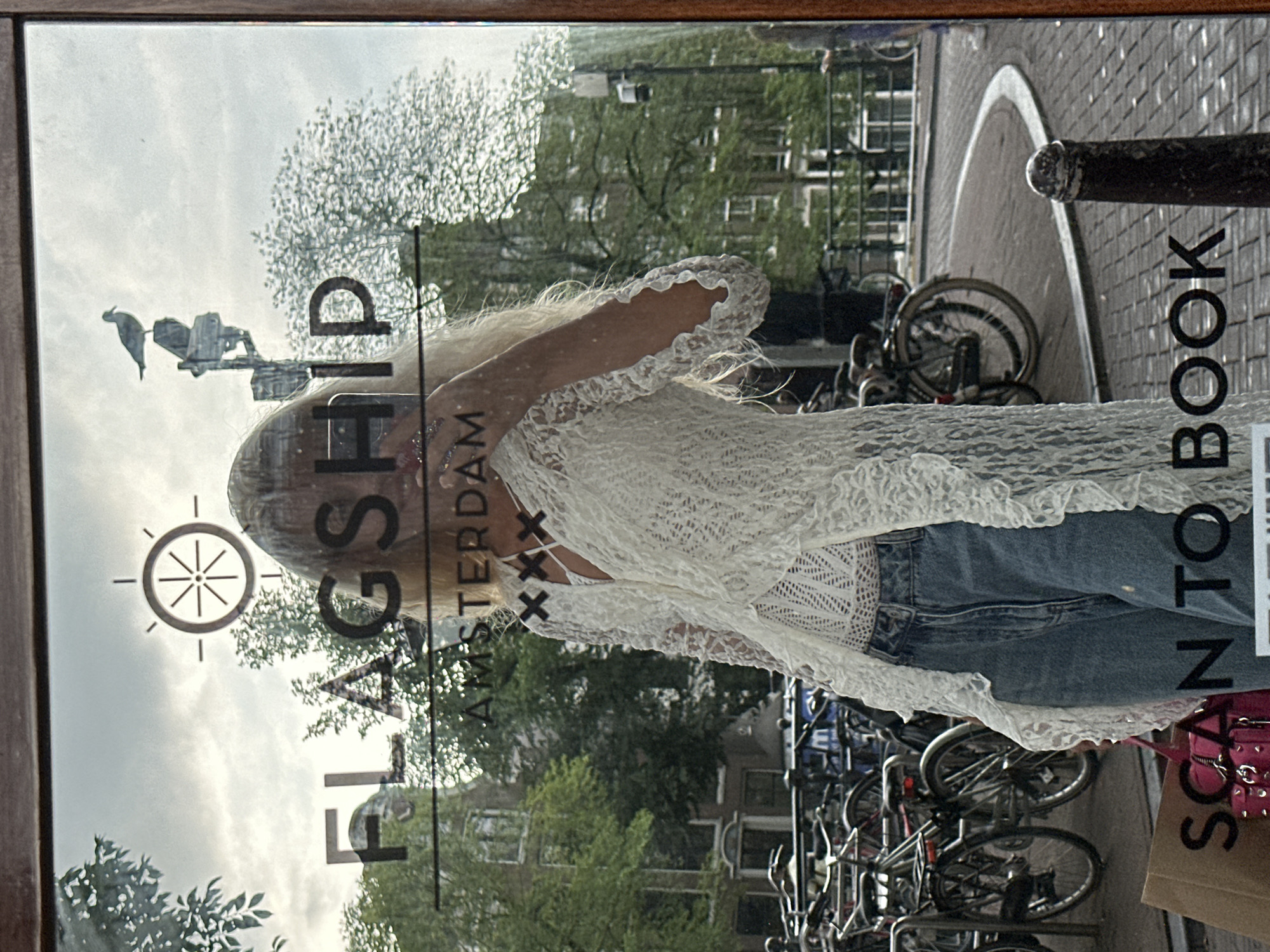 A young woman with blonde hair is standing in front of a shop window in Amsterdam, Netherlands. She is wearing a white lace top and blue jeans. The shop window has text that reads "FLAGSHIP AMSTERDAM XXX" and "SCAN TO BOOK". In the background, there are bicycles parked and buildings that are typical of Amsterdam. The weather appears to be overcast. The woman is holding her hand to her face and looking into the camera.