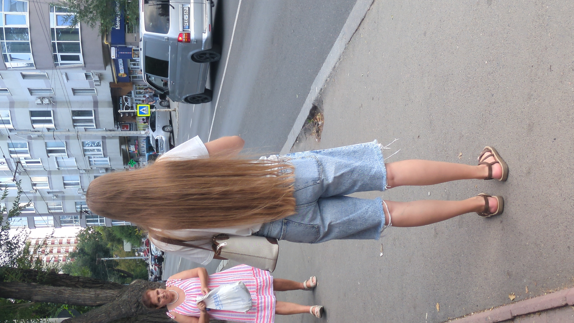 The image shows a street scene in Chișinău, Moldova. In the foreground, a young woman with very long, light brown hair is seen from behind, wearing a white t-shirt and light blue denim shorts with frayed hems. She is also wearing brown sandals. To her left and further back, an older woman with dark hair, wearing a pink and white striped dress, is holding a white plastic bag. She is wearing light-colored sandals. Both women are walking on a paved sidewalk next to a street. Several cars, including a silver van with license plate "FEE 167", are visible on the street. In the background, there is a multi-story building with many windows and balconies, typical of urban architecture in Chișinău. Trees and streetlights are also present. The overall impression is a casual, everyday urban setting.