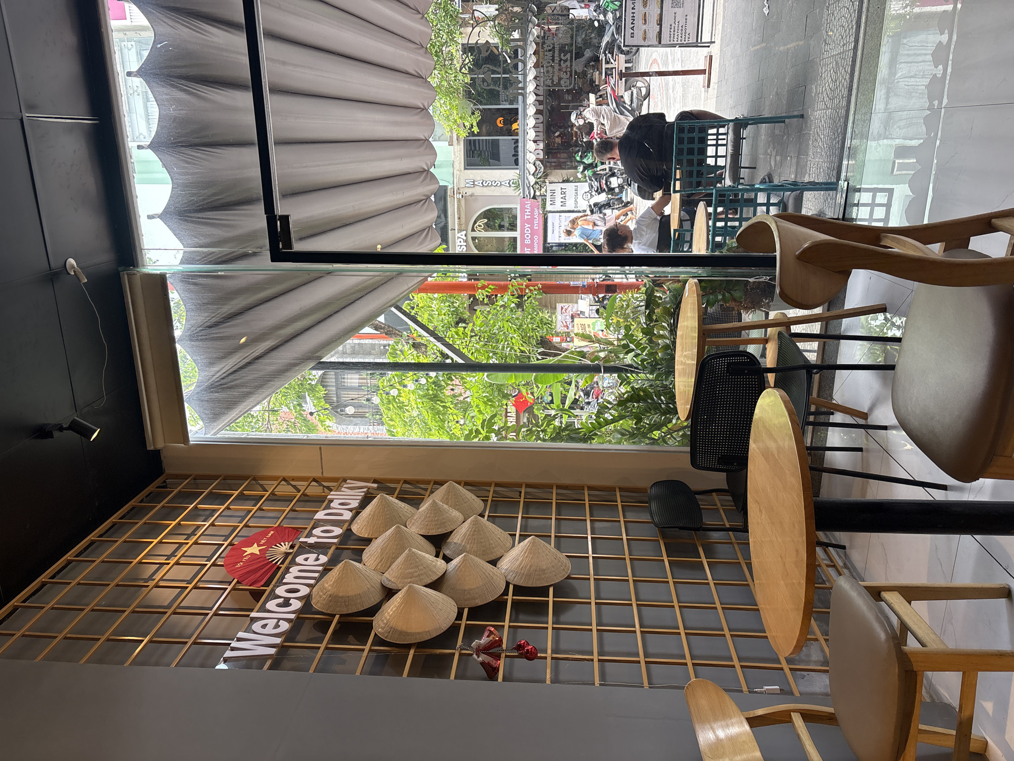 The image shows the interior of a café in Đà Nẵng, Vietnam, next to a window view of a street scene. The café decor includes woven bamboo latticework on the wall, adorned with traditional Vietnamese conical hats, a red fan, and a “Welcome to Dalky” sign. Tables and chairs are arranged inside. Outside the café, the street is visible through the window, with pedestrians, scooters, and shops including a spa, massage parlor, and mini-mart. The street scene is partially obscured by a retractable awning.