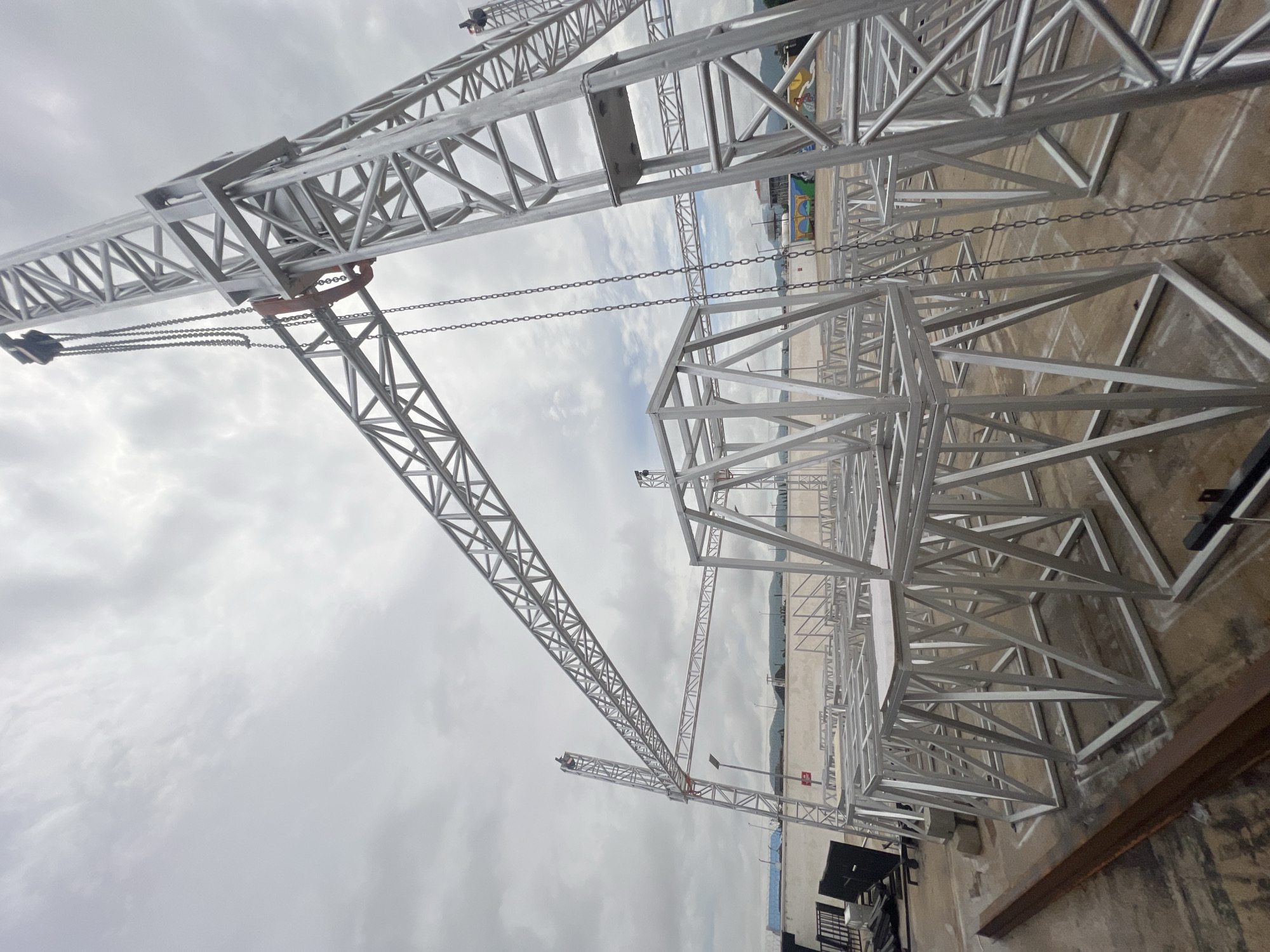 The image displays a construction site in Howayi, Nigeria, with a focus on scaffolding and cranes. The metallic structures are the primary subjects, featuring intricate lattice designs typical of industrial equipment. Chains hang from some of the cranes, suggesting ongoing assembly or preparation for lifting heavy materials. The scene is set outdoors, with a concrete or asphalt ground visible. In the background, a body of water is discernible, bordered by a distant shoreline with hints of buildings and possibly some colorful structures, suggesting a coastal or waterfront location. The sky is overcast with diffused light, indicating either an early morning, late afternoon, or a day with heavy cloud cover. There are no people prominently visible in the frame, implying the activity might be paused or focused on the machinery itself. The overall atmosphere is one of industrial activity and construction, with a sense of scale conveyed by the towering metal structures. The location context, Howayi, Nigeria, further grounds the image in a specific geographical setting.