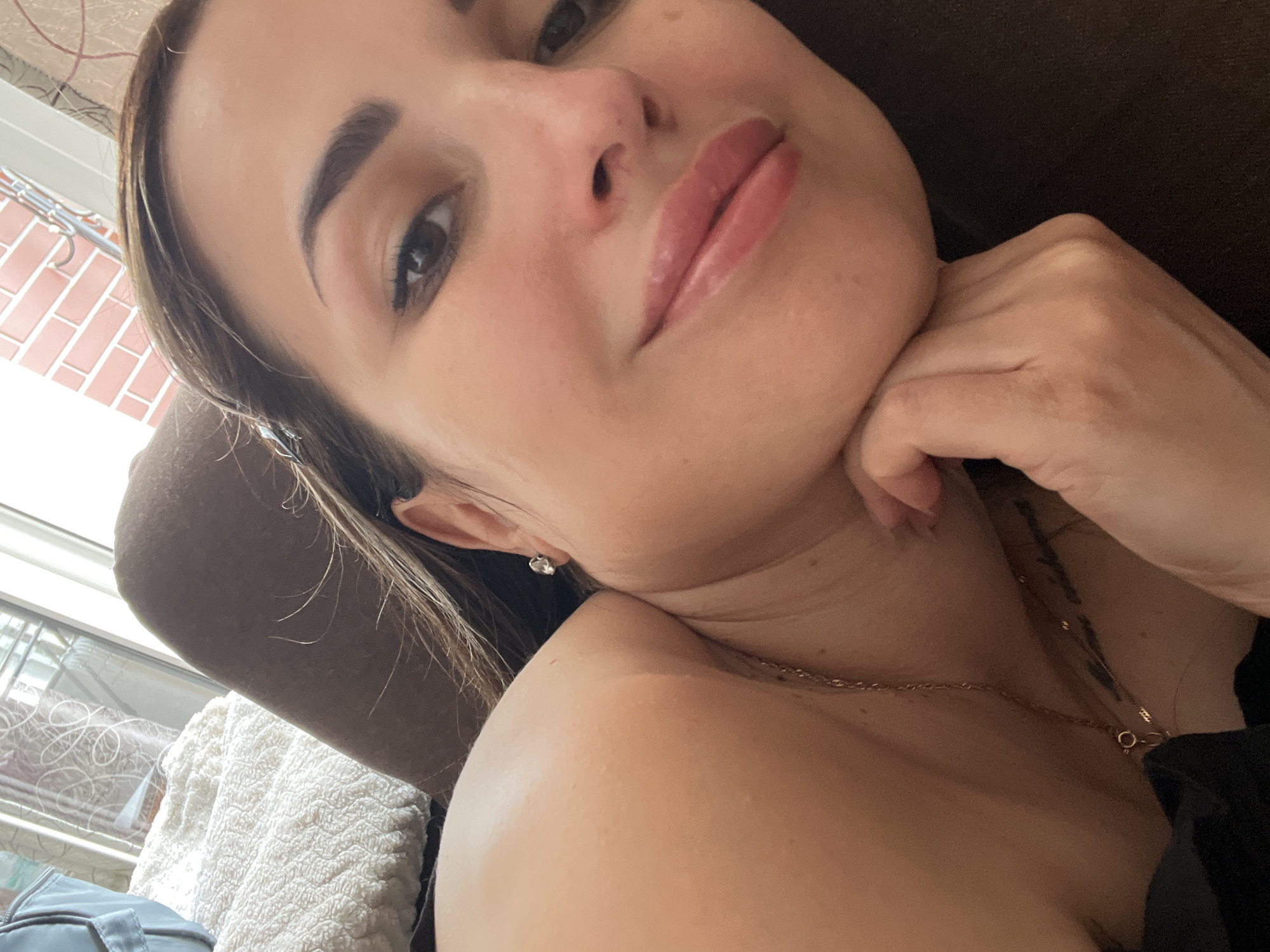 A close-up, over-the-shoulder selfie shows a woman with long, brown hair, dark brown eyes, and full lips. She is lying down with her head resting on her hand, which is propped on her chin. She is wearing a delicate gold necklace. In the background, a textured white blanket is visible, along with a glimpse of a window with white curtains and a hint of red brick. The lighting suggests an indoor environment, possibly a bedroom or living room.