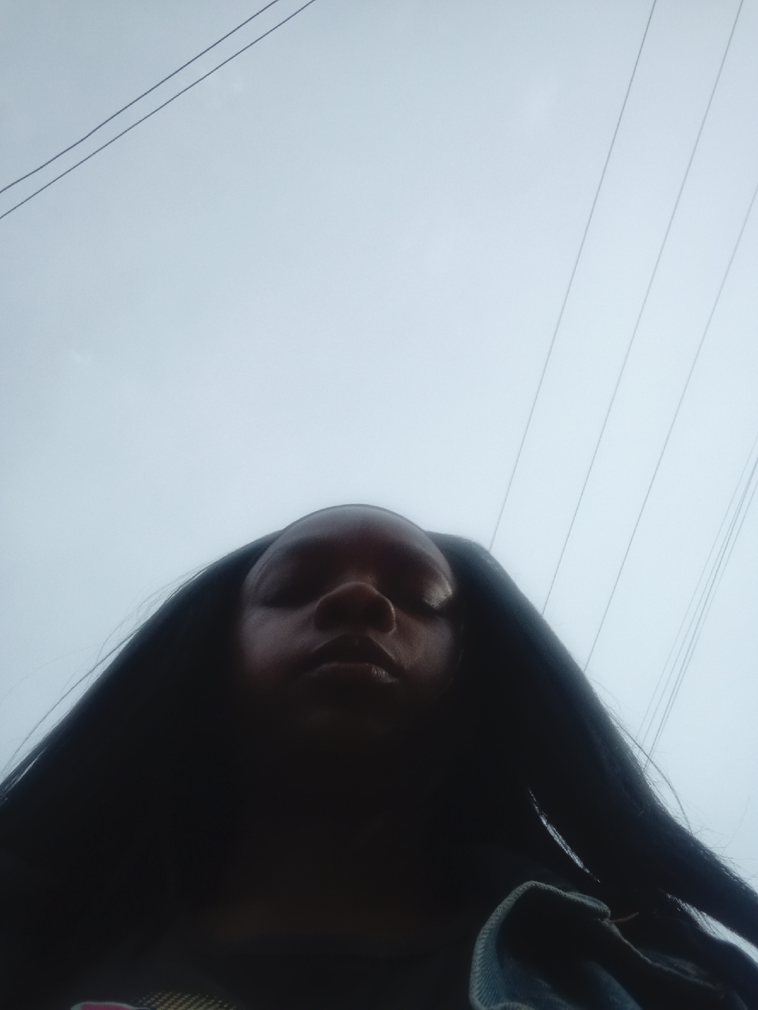 The image shows a low-angle view of a person with dark hair and skin, facing the camera with closed eyes. Above the person is a light grey sky with power lines visible in the upper portion of the frame. It is difficult to determine the location in Mahiakalo ward, Kenya, because there are no distinguishable landmarks. The focus is on the person, with the sky and power lines serving as the background. The presence of power lines suggests an urban environment.