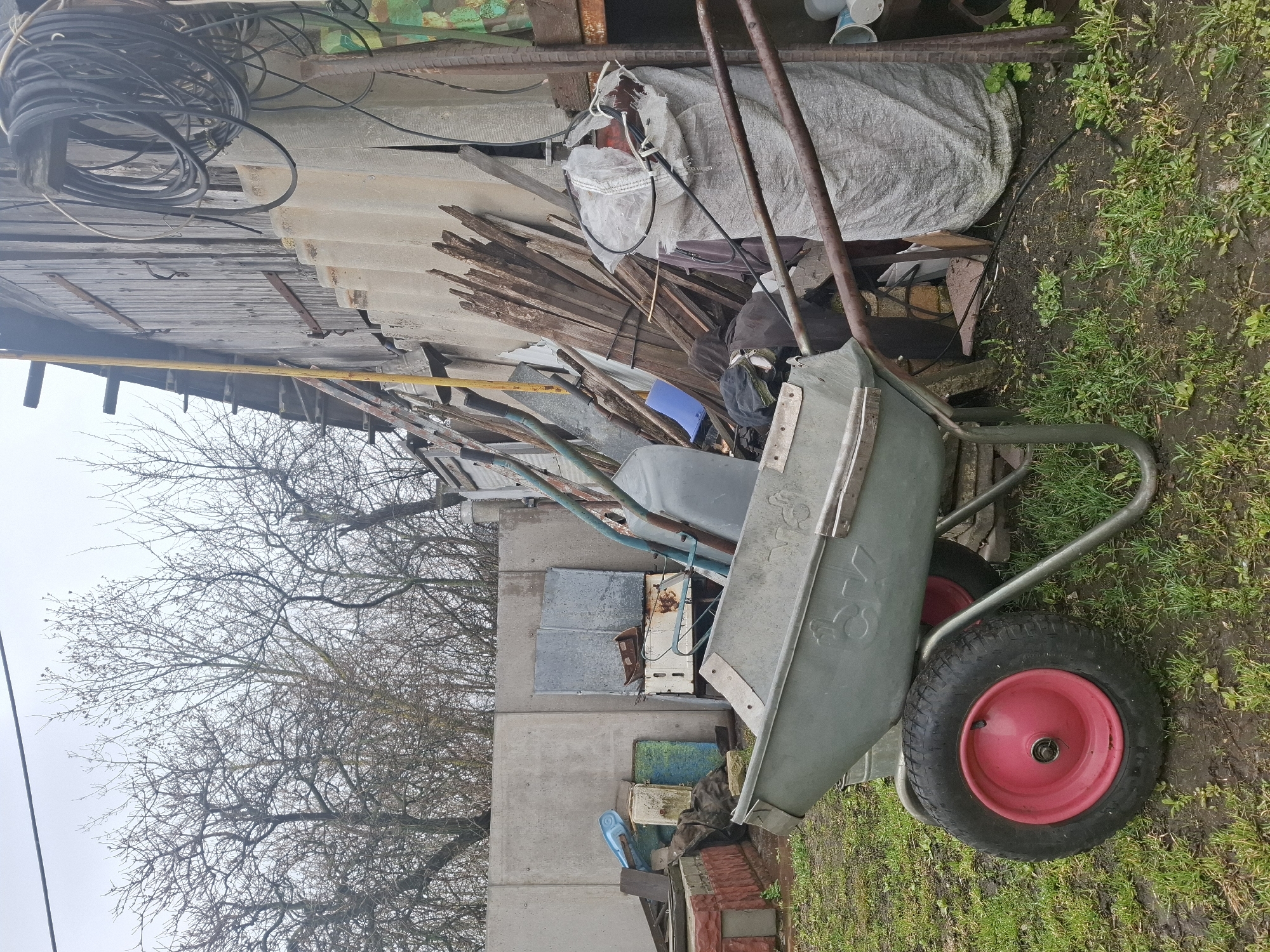 The image depicts a cluttered outdoor scene in what appears to be a yard or storage area, likely located in a rural or semi-rural setting. A light-colored wheelbarrow with a red wheel is prominently displayed in the foreground, situated on damp grass. Behind the wheelbarrow is a disorganized collection of materials including lumber, corrugated roofing sheets, metal scraps, and large bags, suggesting the presence of construction or demolition debris. A section of a weathered wooden structure is visible on the left side of the frame. In the background, a leafless tree stands against a grey sky, indicating a cold or off-season time of year. The overall impression is one of disarray and accumulation of materials. The location, Khlevnoye, Russia, is mentioned. There are no people present in the image.