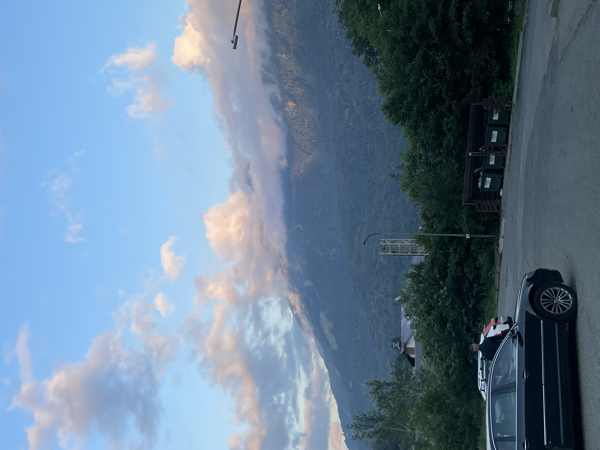 The image captures a scenic view of the mountains surrounding Krasnaya Polyana, Russia, under a partly cloudy sky with soft pink and orange hues from the setting sun. In the foreground, a dark-colored sedan is parked on an asphalt surface. Beside it, another car, partially visible, is parked behind a person in a dark shirt. Further in the background, a row of green garbage bins is situated near a dense line of trees. The mountains, covered in lush greenery, form the backdrop, with some peaks partially obscured by fluffy clouds. A tall, industrial-looking structure is also visible among the trees, suggesting a proximity to infrastructure within the natural landscape.
