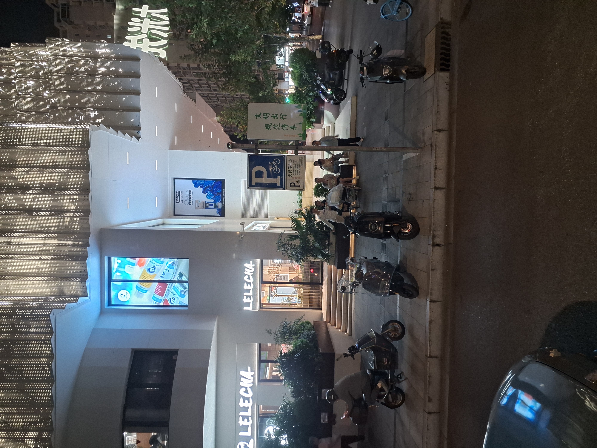 The image depicts a street scene at night in Shanghai, China, likely in a commercial or shopping district. A modern building with a distinctive perforated facade is visible on the left, featuring illuminated signage for "LELECHA". Adjacent to it, another entrance for "LELECHA" is also illuminated. A large digital screen displays colorful paint tubes, possibly advertising art supplies. Several parked electric scooters and motorcycles line the sidewalk. A group of people are seated on a bench, some with a baby stroller. A person on a scooter is looking at their phone. A "P" parking sign with a bicycle icon is posted. Another sign with Chinese characters related to civilized behavior and parking is visible. Trees and more buildings are present in the background. The street is paved, and a car is partially visible in the foreground. The lighting suggests it is late evening or night.