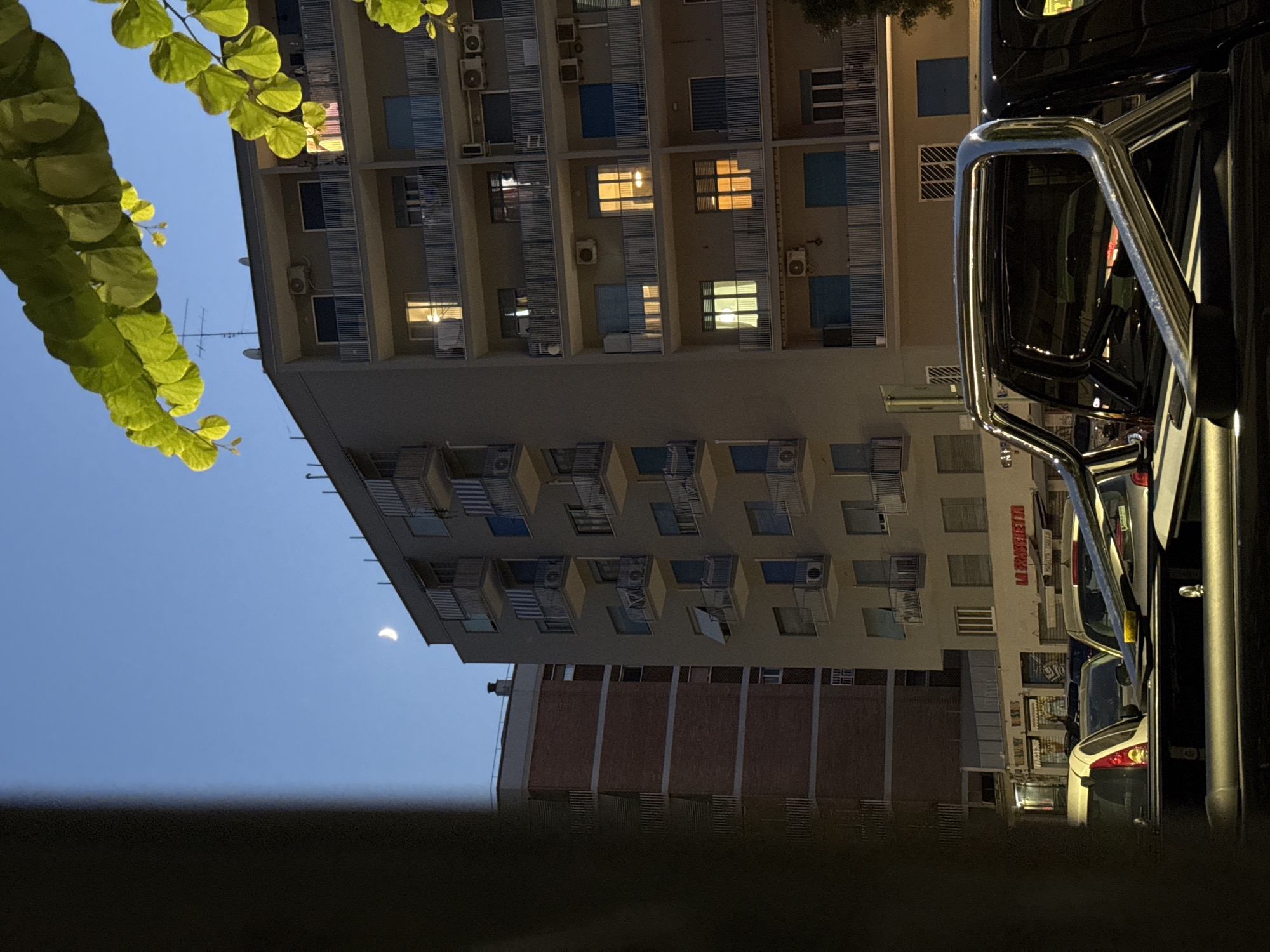 The image shows a street scene in Rome, Italy, at dusk. In the foreground, the back of a black pickup truck with a chrome roll bar is visible. Beyond the truck, several cars are parked on the street. In the background, there are apartment buildings. The building directly behind the cars appears to be a lighter color with balconies and windows, some of which are lit. To the left of this building, there is another building made of red brick. Above the buildings, the sky is a deep blue with a crescent moon visible. Green leaves from a tree are in the upper right corner of the frame, partially obscuring the sky and building.