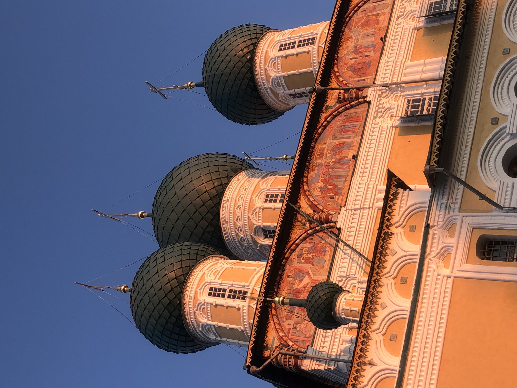 A low-angle, close-up outdoor shot captures the upper portion of the Resurrection Cathedral in Tutaev, Russia, during what appears to be sunset or sunrise. The cathedral's architecture is characterized by multiple onion domes, painted murals, and decorative white trim against an orange facade. Three large, dark green onion domes with diamond-shaped scale patterns are prominently featured, each topped with a cross. Smaller, dark green domes are visible as well, including one that juts out in the foreground, also crowned with a cross. The domes are supported by ornate cylindrical towers with arched windows and detailed white moldings. Below the domes, a band of colorful murals depicts religious scenes with figures in robes. The sky is a clear, deep blue. The warm, golden light of the low sun illuminates the right side of the building, highlighting the architectural details and casting soft shadows. The image focuses on the grandeur and decorative elements of the cathedral, with no people or contemporary objects visible.