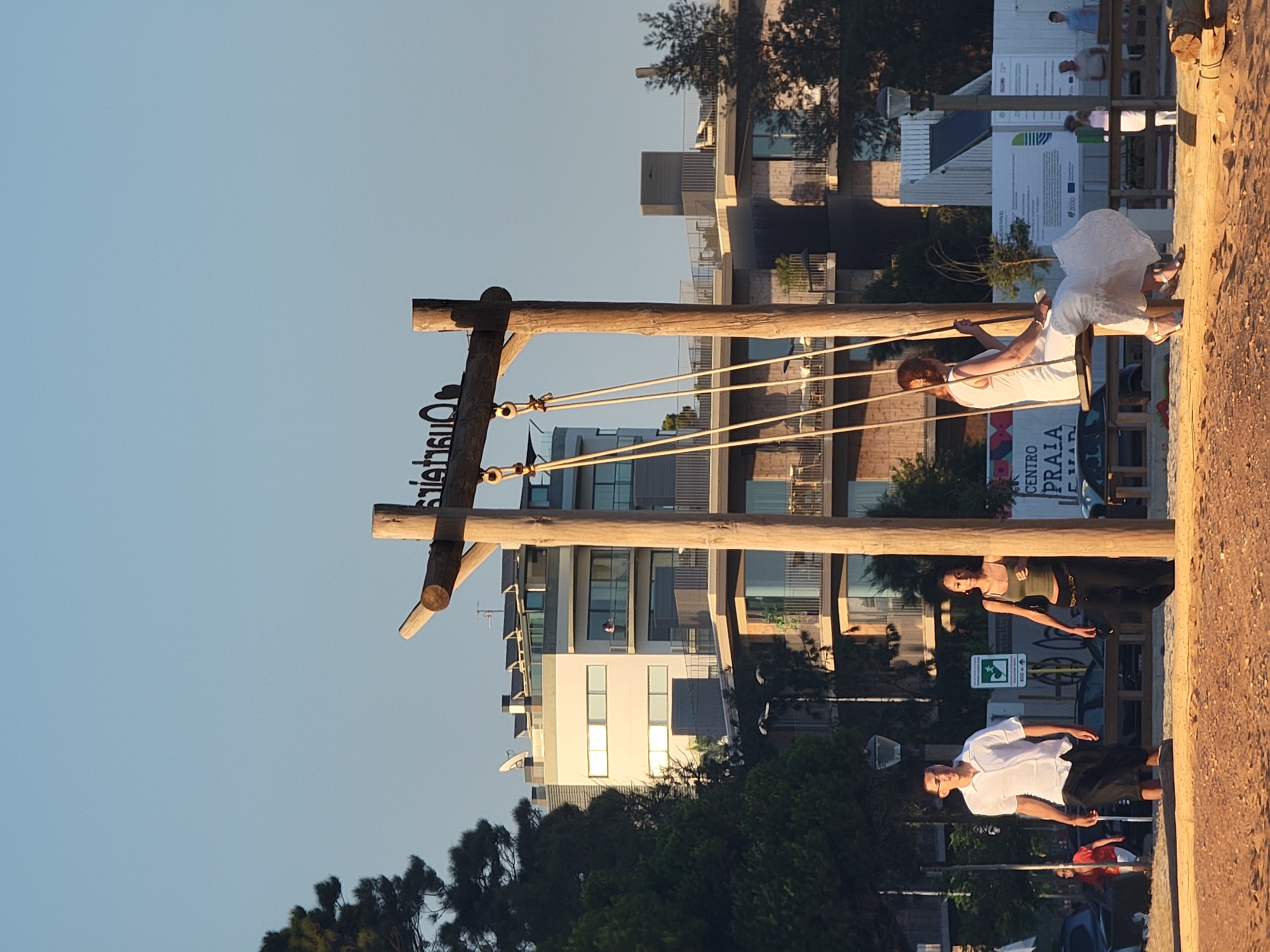 The media file shows a beach scene in Quarteira, Portugal, likely in the late afternoon or early evening due to the warm, golden light. The sky is clear and a pale blue. The main subjects are several people engaged in various activities near a large wooden swing structure.
In the foreground, a person in a white top and dark shorts is standing, appearing to be looking towards the left. Behind them, a person wearing a green top and dark pants is standing on the sand, also looking to the left, with their arm extended. Further back, a person in a white dress is seated on a swing, holding a phone in their hand. They are positioned on the swing, suggesting they are about to swing or have just finished.
The setting includes a beach with sandy ground and a wooden structure that appears to be part of a playground or recreational area, featuring ropes and wooden beams. In the background, there are modern buildings with balconies and large windows, characteristic of a coastal resort town. Some greenery, including trees, is visible in the mid-ground and background.
A notable detail is a sign with text that appears to be in Portuguese. Some visible words include "CENTRO," "PRAIA," and "ALGARVE." There is also a white sign with a green symbol that might be a directional or informational marker.
The overall mood seems relaxed and leisurely, consistent with a day at the beach. The presence of people on the swing and standing around suggests a social gathering or a tourist activity. The lighting indicates it is either sunrise or sunset.