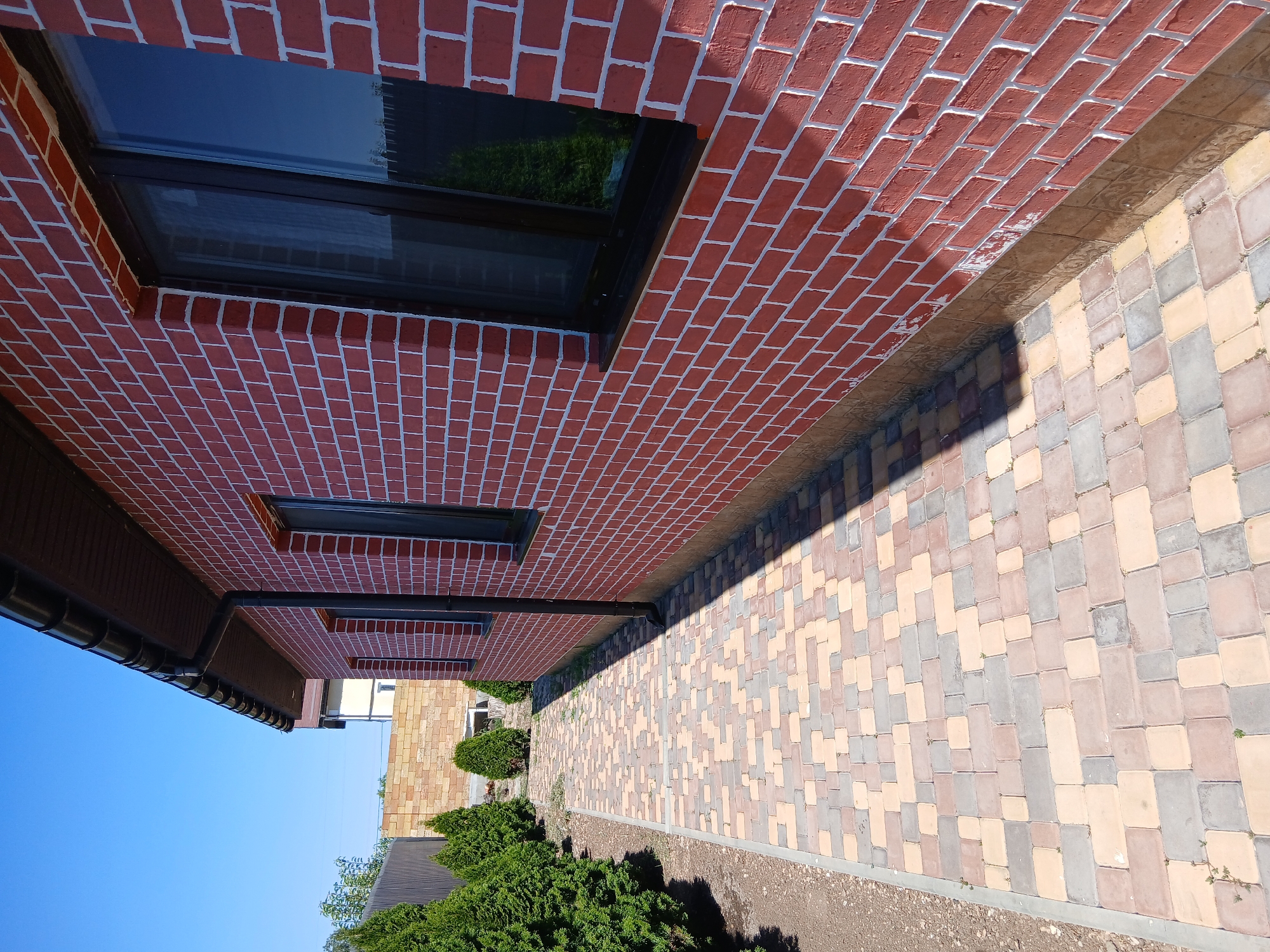 This image shows a side view of a red brick house with white mortar, featuring dark-framed windows. A paved walkway, made of multi-colored rectangular stones, runs alongside the house. Green conifer trees are planted along the edge of the walkway. In the background, a fence and another building can be seen. The sky is clear blue. The scene is located in the city of Mykhailivka, Ukraine.