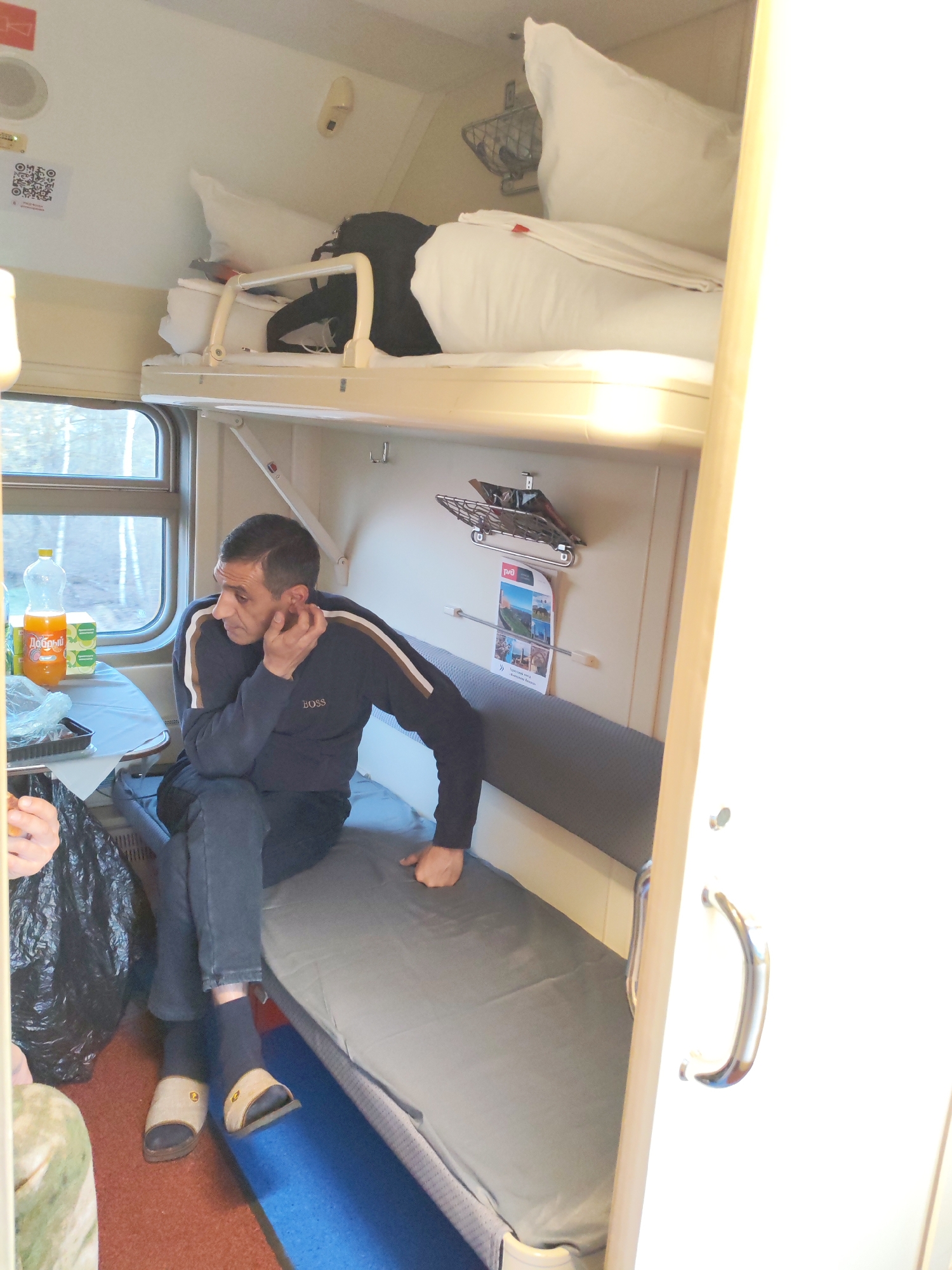 This media file captures the interior of a train compartment, likely a Russian sleeper car (platscard or kupe). The scene is brightly lit by natural light streaming through a large window on the left, suggesting daytime.
The primary subject is a man sitting on the lower bunk bed on the right side of the compartment. He is wearing a dark blue long-sleeved sweatshirt with "BOSS" visible on the chest, dark trousers, and light-colored slippers with a gold emblem. He is looking towards the window, with his right hand near his ear in a thoughtful or casual pose. His expression is neutral.
The compartment features two bunk beds. The upper bunk, visible at the top, is made with white bedding, pillows, and a duvet, with a safety rail. Some dark clothing or a bag is stored on the upper bunk. Above the window, a small overhead rack is present. On the small table next to the window, an orange plastic bottle of "Добрый" (Dobry), a Russian juice brand, and a carton of juice are visible. A black plastic bag is on the floor near the table.
On the wall above the man's head, there's a small metal rack with a poster or leaflet hanging from it, displaying scenic images and a "РЖД" (RZD - Russian Railways) logo. Further to the left on the wall, near an overhead speaker, a white QR code sticker is affixed. Other details include coat hooks and a light fixture. A partial view of another person's hand and leg is visible in the lower-left corner, suggesting another passenger. The floor is covered with a reddish-brown carpet and a blue mat.
