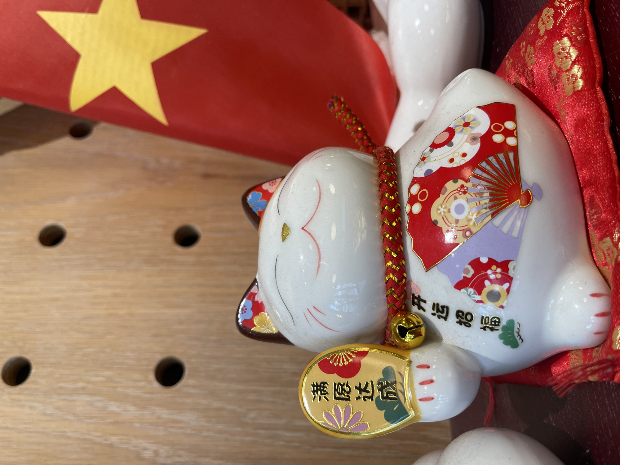 The image displays a white ceramic Maneki-neko, or beckoning cat figurine, resting on its back on a red cushion with gold patterns. The cat is adorned with red and gold decorative elements, including floral designs and fans on its body. A red braided cord with a small golden bell hangs around its neck. In its left paw, the cat holds a golden oval plaque with Japanese characters.
Behind the Maneki-neko, a Vietnamese flag is partially visible, featuring a prominent yellow five-pointed star on a red background. The flag is positioned towards the upper left of the frame. The objects are arranged on a light-colored wooden surface with visible grain and several small, dark circular holes. In the background, other out-of-focus white ceramic objects are partially discernible.
No specific activity or event is taking place, as it is a still life arrangement of decorative items. The scene appears to be indoors, potentially in a shop or home in Bắc Nha Trang, Vietnam, as indicated by the presence of the Vietnamese flag.
Visible text on the golden plaque held by the Maneki-neko reads "千萬両" (Sen Man Ryō), which symbolizes "ten million ryo" or great wealth. On the cat's body, near its right paw, the characters "開運招福" (Kaiun Shōfuku) are visible, meaning "good luck and good fortune."