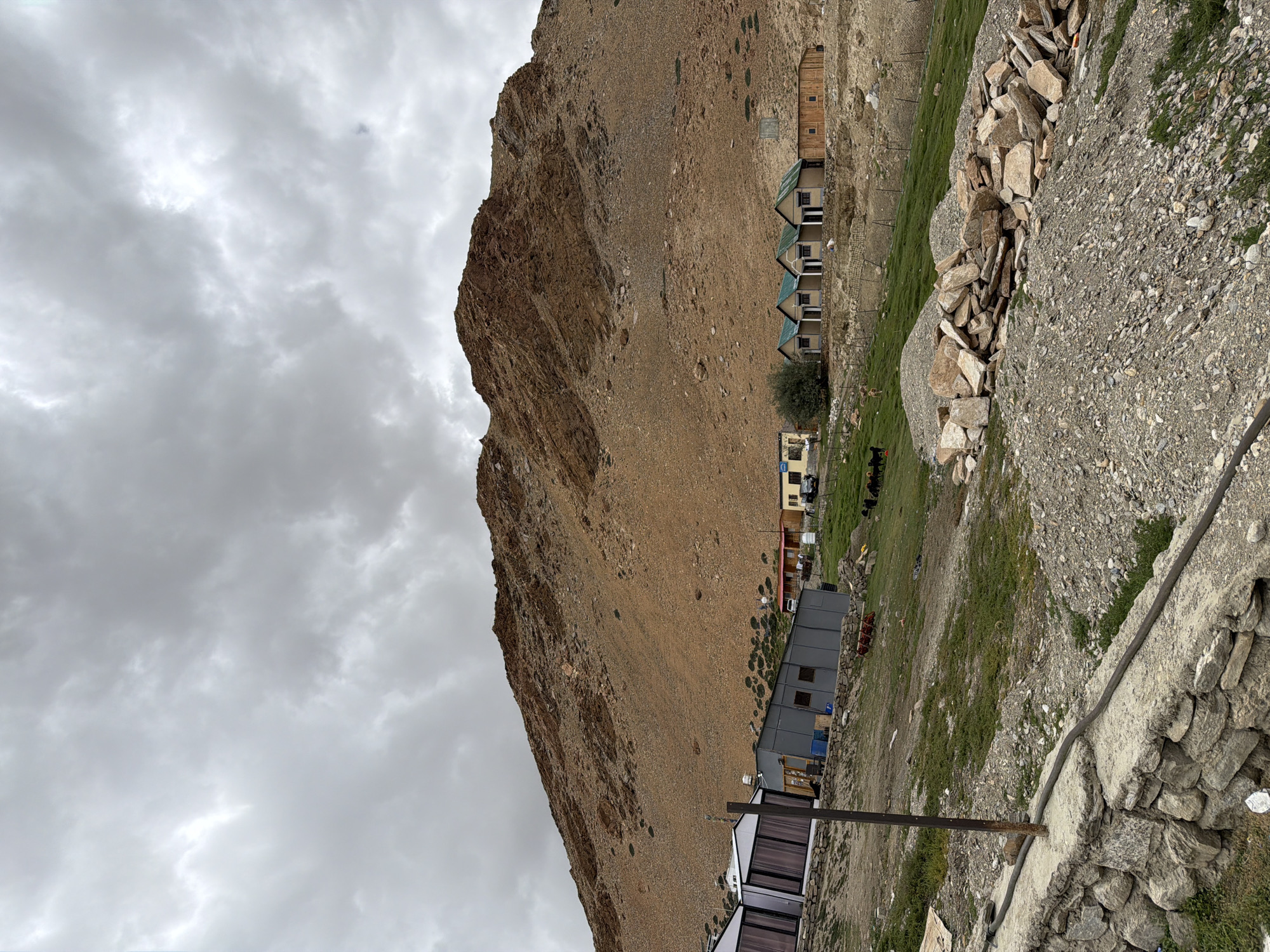 A medium shot captures a village with small houses and structures in front of a large, barren mountain under a cloudy sky. The houses are small, with green roofs, and are lined up in a row. Some of the structures are modern, with glass windows, and others are made of wood. A dirt road runs through the village, and there are a few cars parked. The mountain is covered in brown rocks and sparse vegetation. The sky is overcast with gray clouds, suggesting a cool or possibly rainy day. The scene appears to be a remote village in a mountainous region. This location is likely in the village of Karzok, India, which is known for its high-altitude setting and proximity to the Himalayas.