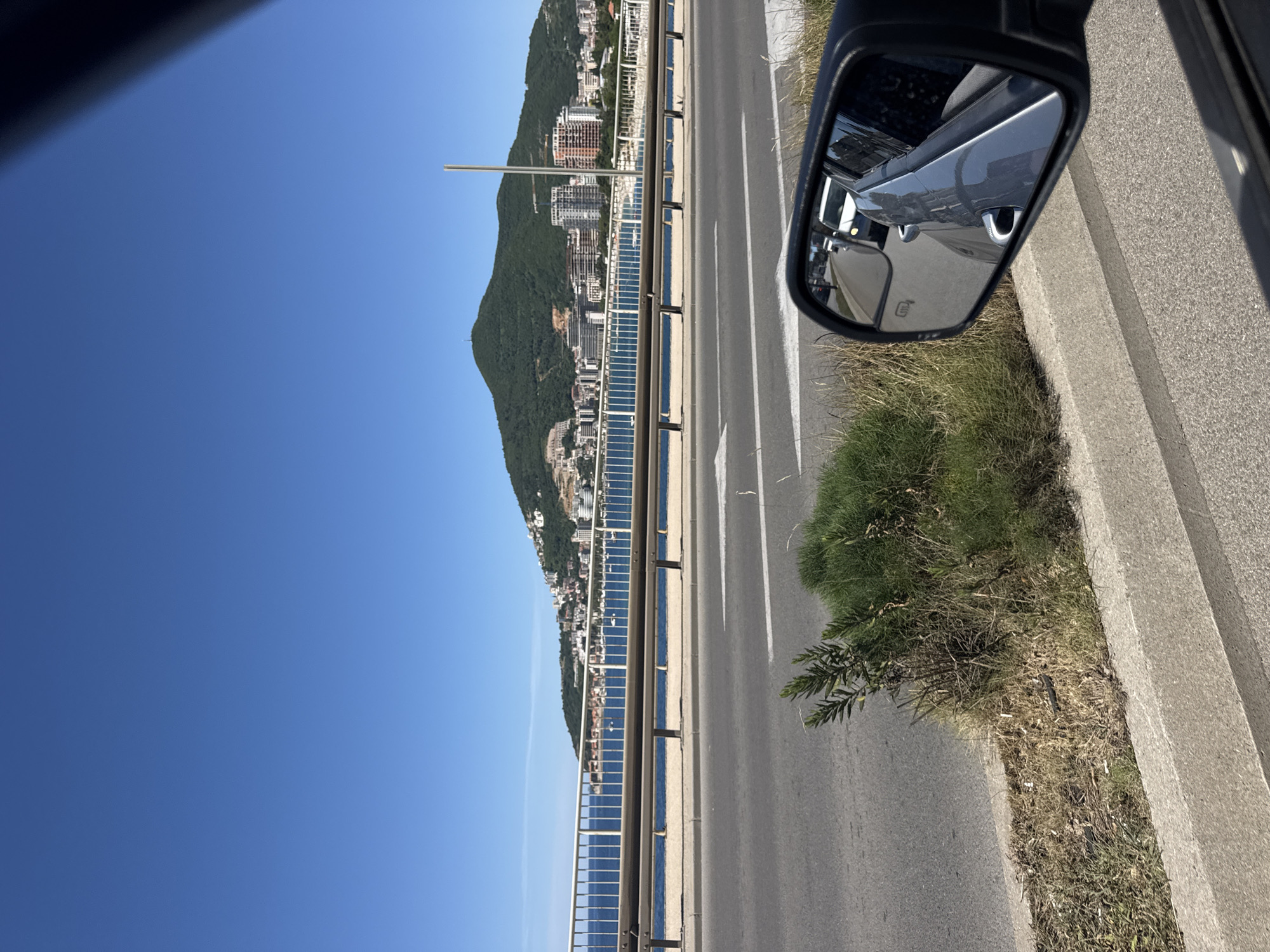 A view from a car on a road in Budva, Montenegro. The road has white lines marking lanes, with a guardrail separating it from the Adriatic Sea. Buildings, likely apartments and hotels, line the coast on the other side of the guardrail. A green mountain rises behind the buildings. The car's side mirror is visible in the upper right, reflecting cars behind it. Grass and weeds grow along the roadside. The sky is clear and blue.