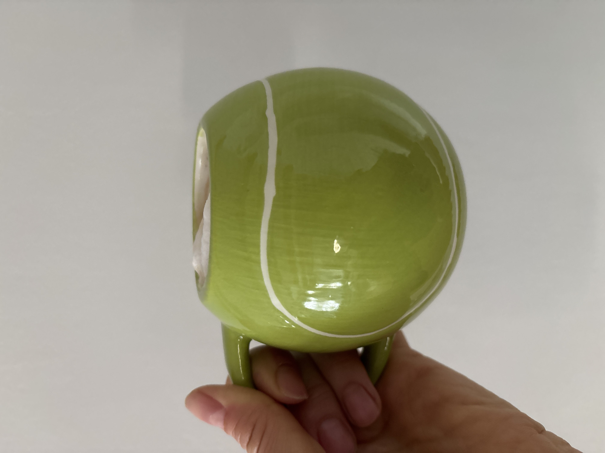 The image displays a close-up, eye-level view of an adult human hand holding a spherical, lime-green ceramic mug against a plain, light grey background. The mug is designed to resemble a tennis ball, featuring two prominent white, curved lines on its exterior and a matching green handle. It contains a light beige liquid topped with white foam. The hand, exhibiting light skin tone and manicured fingernails, securely grips the mug's handle. No other objects or environmental details are visible. This scene is situated in Chaikovsky, Russia.