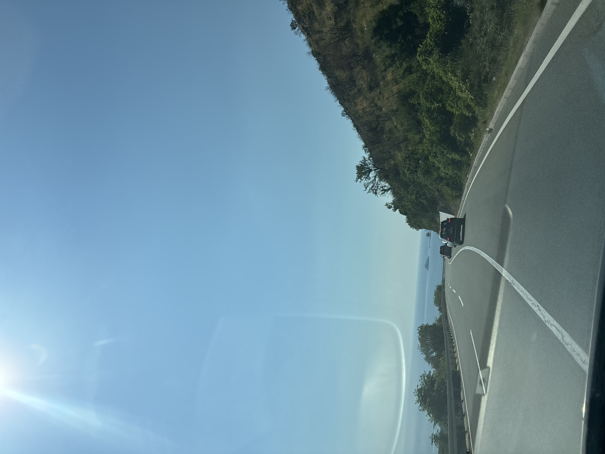 The image depicts a highway in Kanjoši, Montenegro, likely taken from a vehicle moving along the road. The scene shows a winding asphalt road with white lane markings, bordered by a metal guardrail on the right. To the left of the road, a steep, tree-covered hillside rises, with sparse vegetation and rocky patches visible. In the distance, the sea or a large body of water is visible, with a hazy horizon. Several cars are traveling on the highway in the same direction as the camera. The sky is a clear, bright blue, suggesting a sunny day. The sun is visible in the upper left corner of the image, casting a bright glare and lens flare that obscures part of the sky and the hillside. The overall impression is of a scenic drive along a coastal highway. No text is visible in the image.
