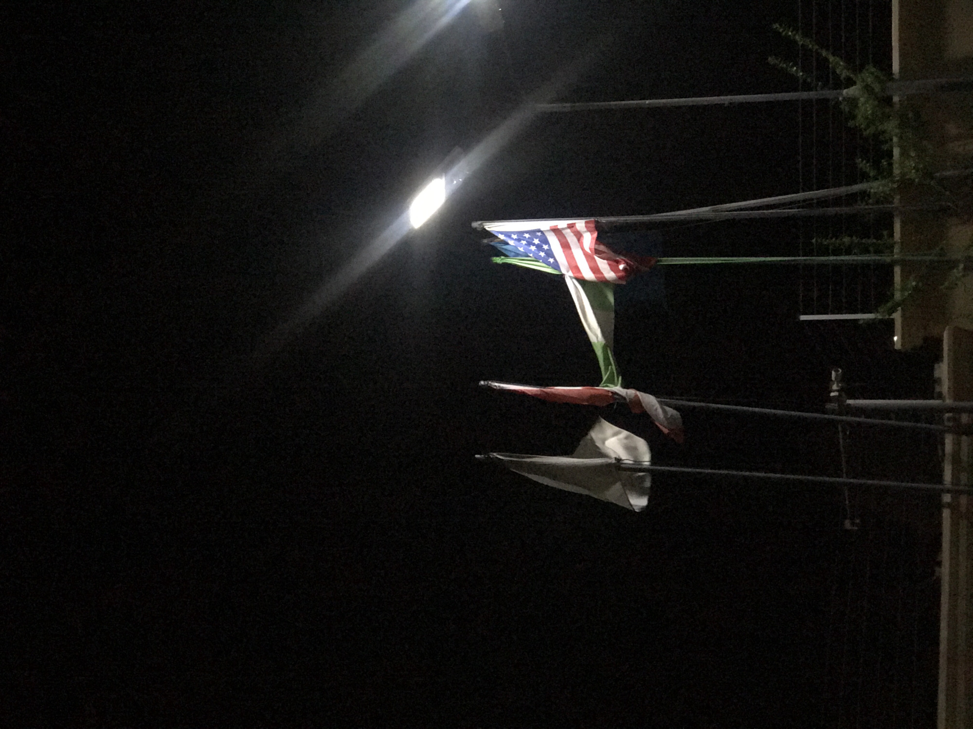 At night, an American flag is affixed to a horizontal pole above an Italian flag. Both flags are strung on horizontal poles and are being illuminated by a bright street light. The flags appear to be somewhere in Abuja, Nigeria.