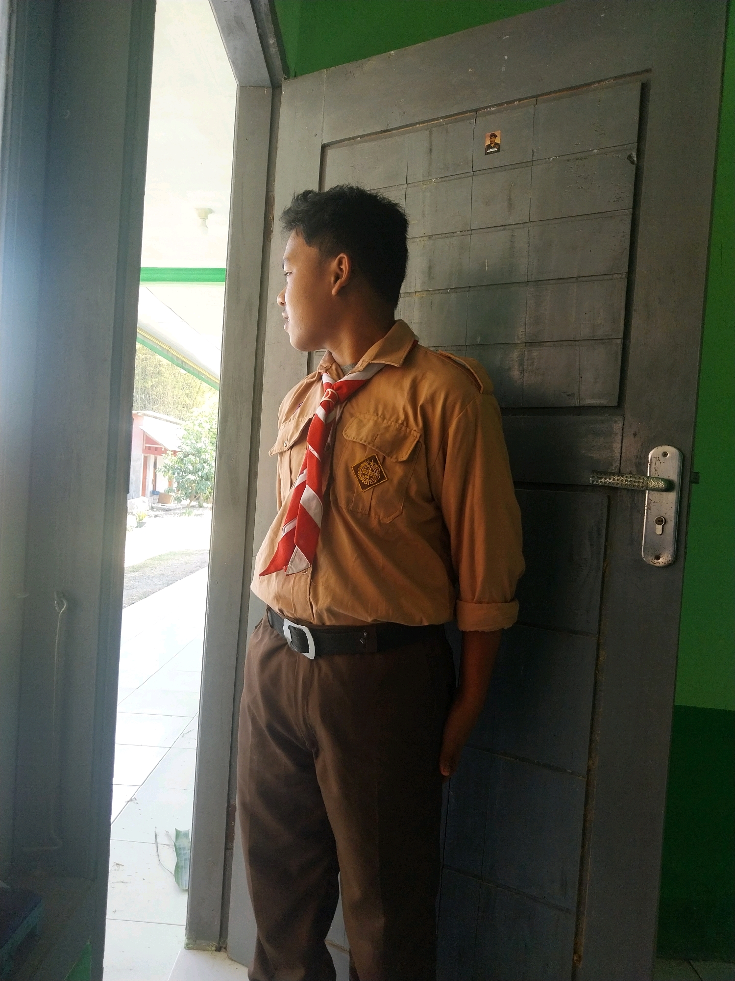 A young person wearing a tan uniform with a red and white striped scarf stands by a doorway, looking to the left. The uniform includes a short-sleeved shirt with a pocket on the left chest bearing a circular emblem, and brown trousers. They are also wearing a black belt with a silver buckle. The doorway is framed by a grey door with visible panels and a silver door handle. To the left of the person, a bright outdoor scene is visible through an open doorway, showing a pathway, some buildings, and greenery. The background to the right of the door is painted green. The image appears to be taken in a school or institutional setting in Pandean, Indonesia, given the uniform and architecture.
