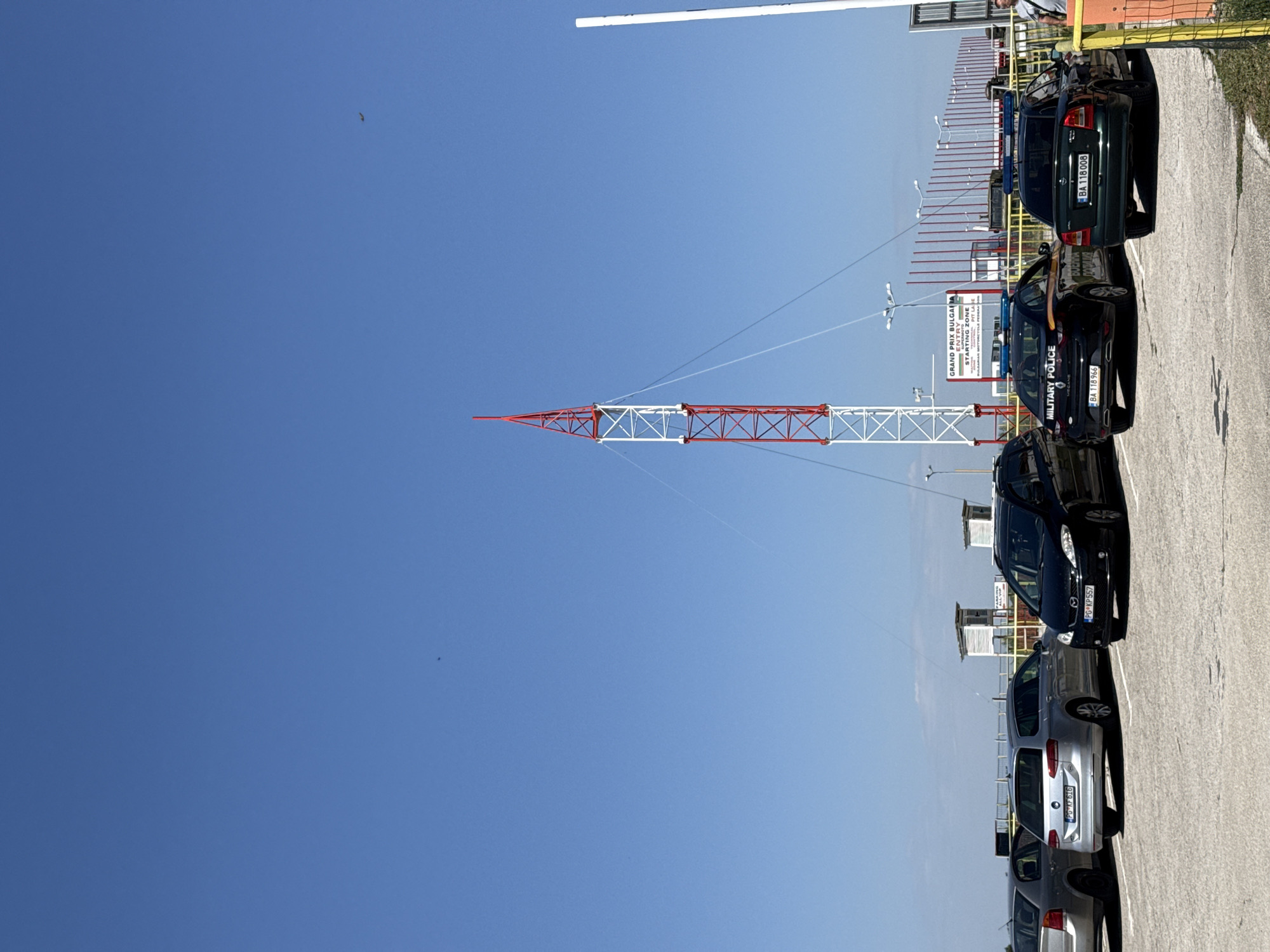 In the city of Dolna Mitropolia, Bulgaria, on a bright, clear day with a vibrant blue sky, a scene unfolds in what appears to be a parking lot or an area adjacent to a facility. Dominating the background is a tall, lattice-style tower, predominantly white with red diagonal accents, a common sight for broadcasting or communication purposes. The tower is anchored by thin wires that extend outwards. To the right, a simpler white pole stands vertically.
In the foreground, several cars are parked. Closest to the viewer are two dark-colored vehicles, one black and one dark green, both identified as "MILITARY POLICE" vehicles by markings on their sides. The black car has a prominent license plate "BA 118 966" and "MILITARY POLICE" written in yellow text. The dark green car displays the license plate "BA 118 008".
Further to the left, a silver station wagon is parked, its license plate visible as "PG KF 616". Another silver car is partially visible to its left, and a darker car is on the far left. Behind the parked cars, a fence with red poles runs horizontally, and beyond that, there are some small structures that appear to be enclosed booths or shelters, one of which has signage reading "GRAND PRIX BULGARIA" and "ENTRY STARTING ZONE". A few indistinct figures can be seen near the fence and the structures. The ground is paved with a light-colored, rough-textured surface, likely asphalt or concrete. The overall impression is an organized outdoor area, possibly related to an event or an official facility, with a strong police presence.