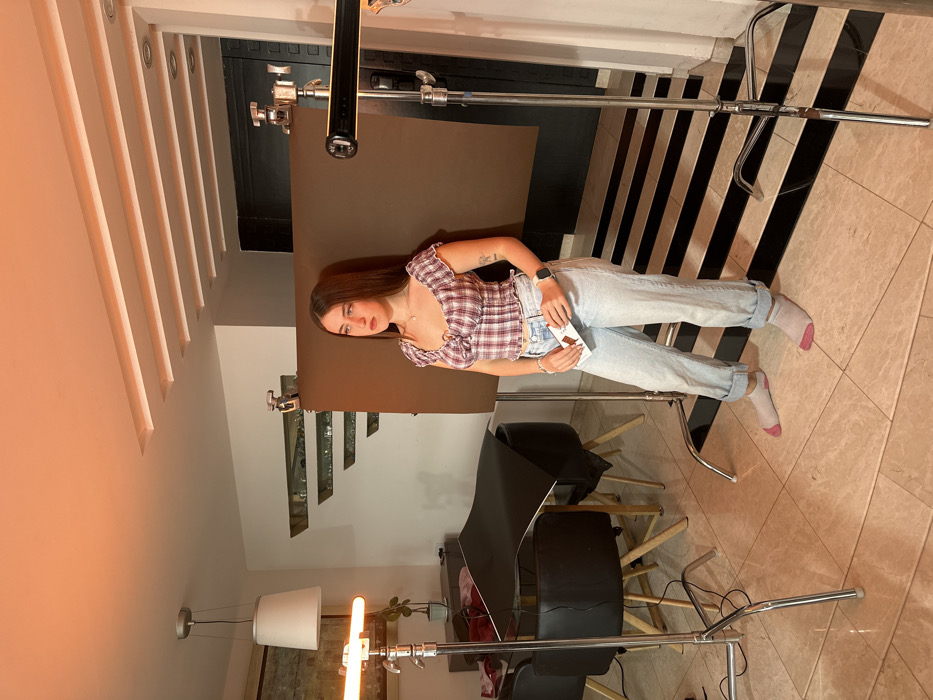 A woman sits with legs outstretched on a tiled floor of black and white stripes, while in the background is a large dark screen propped up by studio equipment. She has long brown hair, wears a pink and white plaid shirt, and light blue jeans with pink socks. She holds a small package in her hand. More lighting and studio equipment are visible, including a black surface with chairs beneath. The room has light colored walls and ceilings with architectural details.