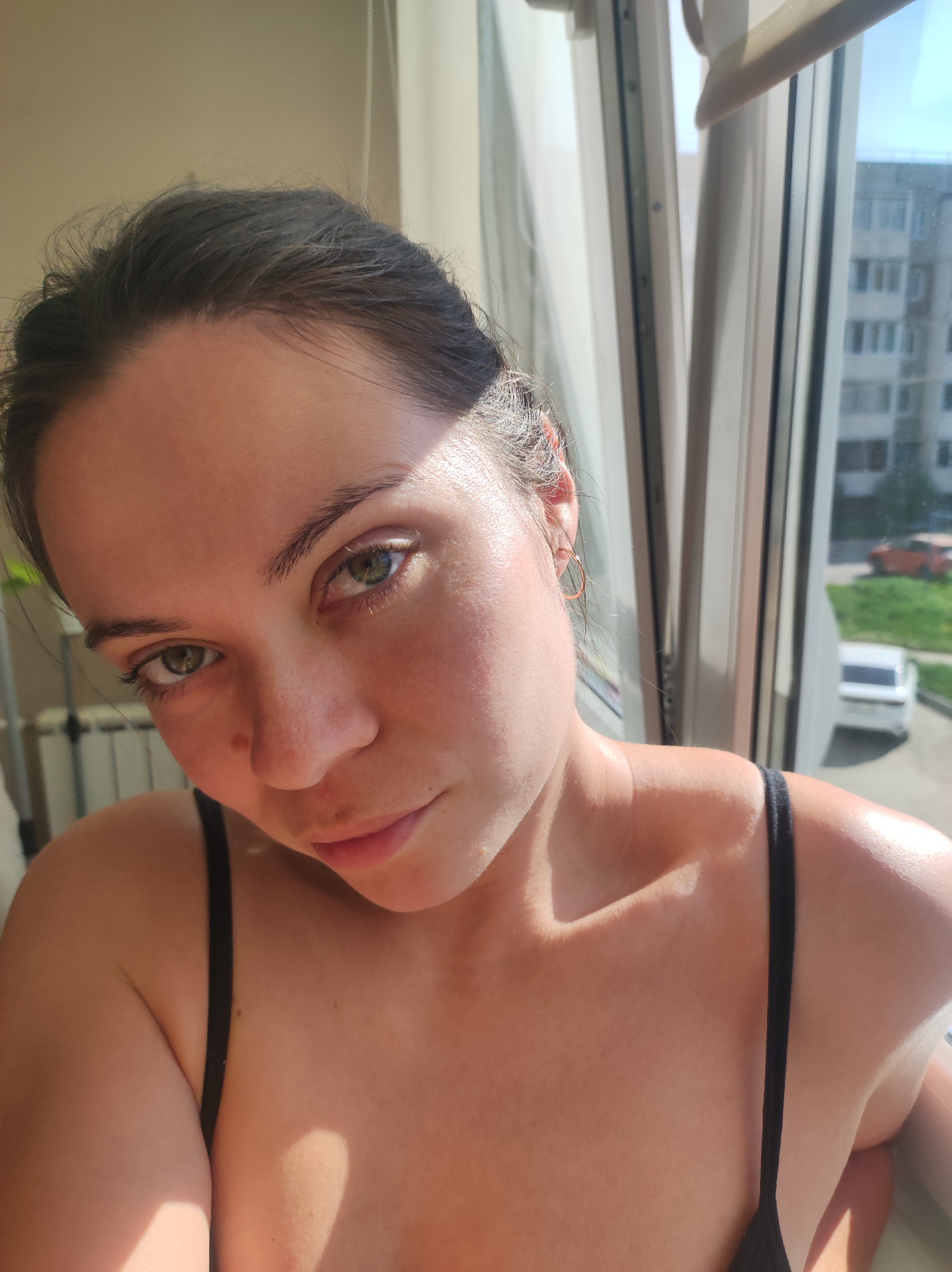 A close-up, high-angle selfie of a light-skinned woman with dark hair pulled back, wearing a black top with spaghetti straps. She has green eyes and is looking directly at the camera. A gold hoop earring is visible in her right ear. Light from a window illuminates the right side of her face and shoulder. The background shows a window with multiple panes and a view of a residential area. In the distance, a multi-story apartment building is visible, and a white car is parked on a street below. Based on the architecture and the type of buildings visible in the background, the location is possibly within the city of Chaikovsky, Russia. A radiator is partially visible to the left.