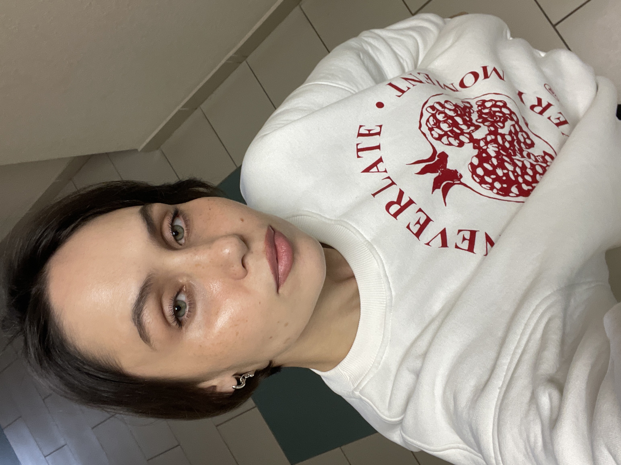 A person with short dark hair and green eyes is lying down, wearing a white sweatshirt with a red design featuring a pomegranate and the words "EVERY MOMENT." The background includes tiled flooring. There is no indication of Izhevsk, Russia or any information to infer the location.