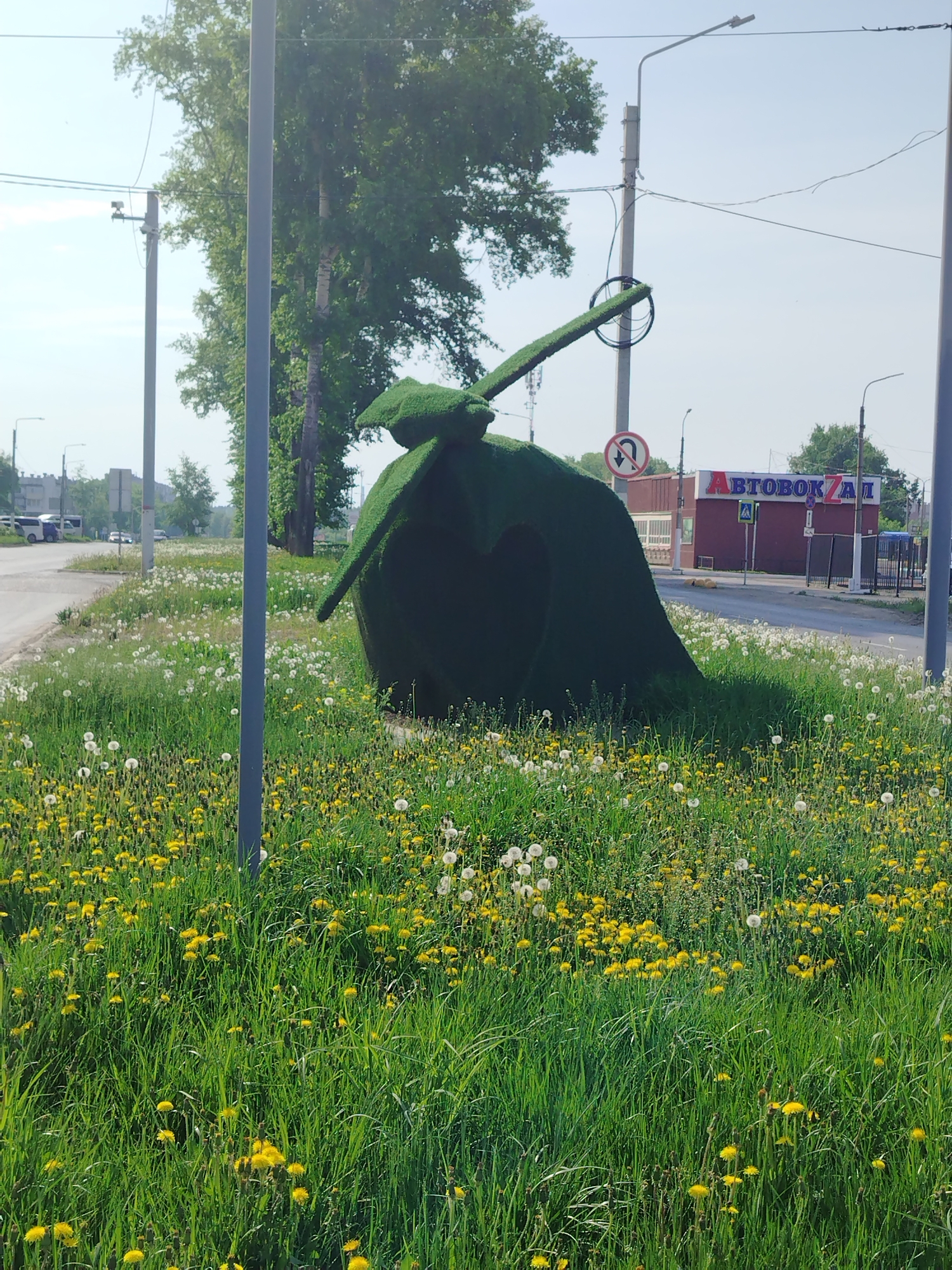 The image shows a green topiary sculpture of a Soviet-era T-34 tank in a grassy area in Chaikovsky, Russia. The tank has a hole in the shape of a heart cut into its side. The grassy area is filled with green grass, yellow dandelions, and white dandelions that have gone to seed. In the background, there is a road with a bus station ("AVTOBOKZAL") sign. The sky is blue and clear. There are lampposts along the road.