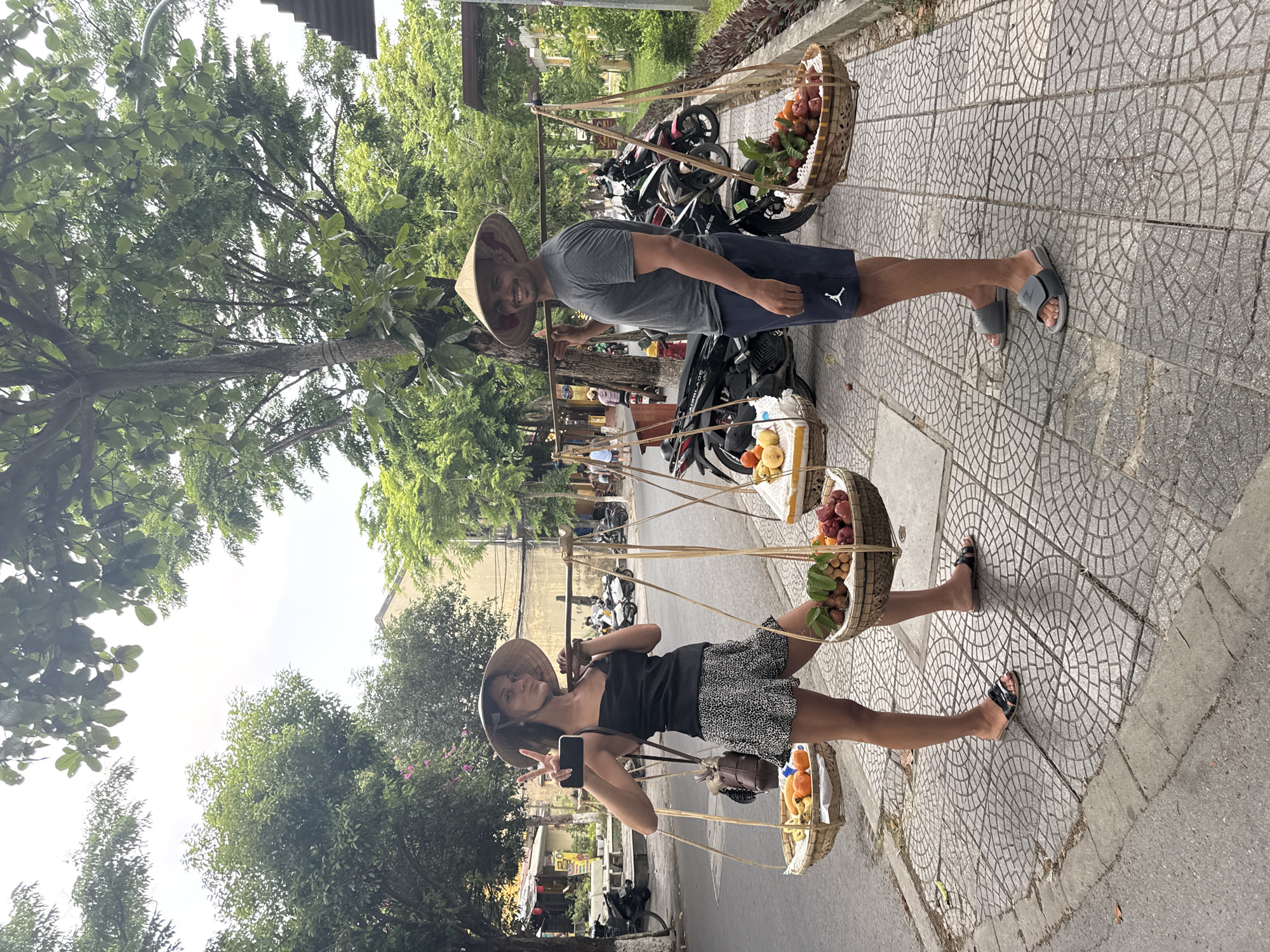 A vibrant outdoor scene in Hội An, Vietnam, captures two individuals, likely tourists, posing with traditional Vietnamese carrying poles (gánh) laden with fresh fruit. The photo is taken on a paved sidewalk, showing a street and lush greenery in the background.
On the right side of the frame, a male stands smiling at the camera. He wears a grey t-shirt, dark shorts with a white "Jordan" logo, and grey slides. A conical straw hat (nón lá) rests on his head, and he holds one end of a bamboo carrying pole from which two woven baskets of fruit are suspended. The upper basket contains red, spiky rambutans, while the lower one holds round, lighter-colored fruits resembling longan or lychees, along with what appear to be oranges or small mangoes.
On the left, a female is also seen smiling, wearing a black tank top, a black and white patterned mini-skirt, and black sandals. She wears a nón lá and holds a smartphone in her left hand while making a peace sign with her right. She also carries a similar gánh with two baskets of fruit, mirroring the male's setup. A small brown purse hangs from her shoulder.
The sidewalk features a distinctive pattern of interlocking circular tiles. In the background, numerous motorbikes are parked along the street, and tall green trees line the area, suggesting a bustling but pleasant urban environment. The scene is brightly lit, indicating daytime, with no harsh shadows, suggesting either an overcast sky or diffused light. Both individuals appear happy and engaged in the photo opportunity.