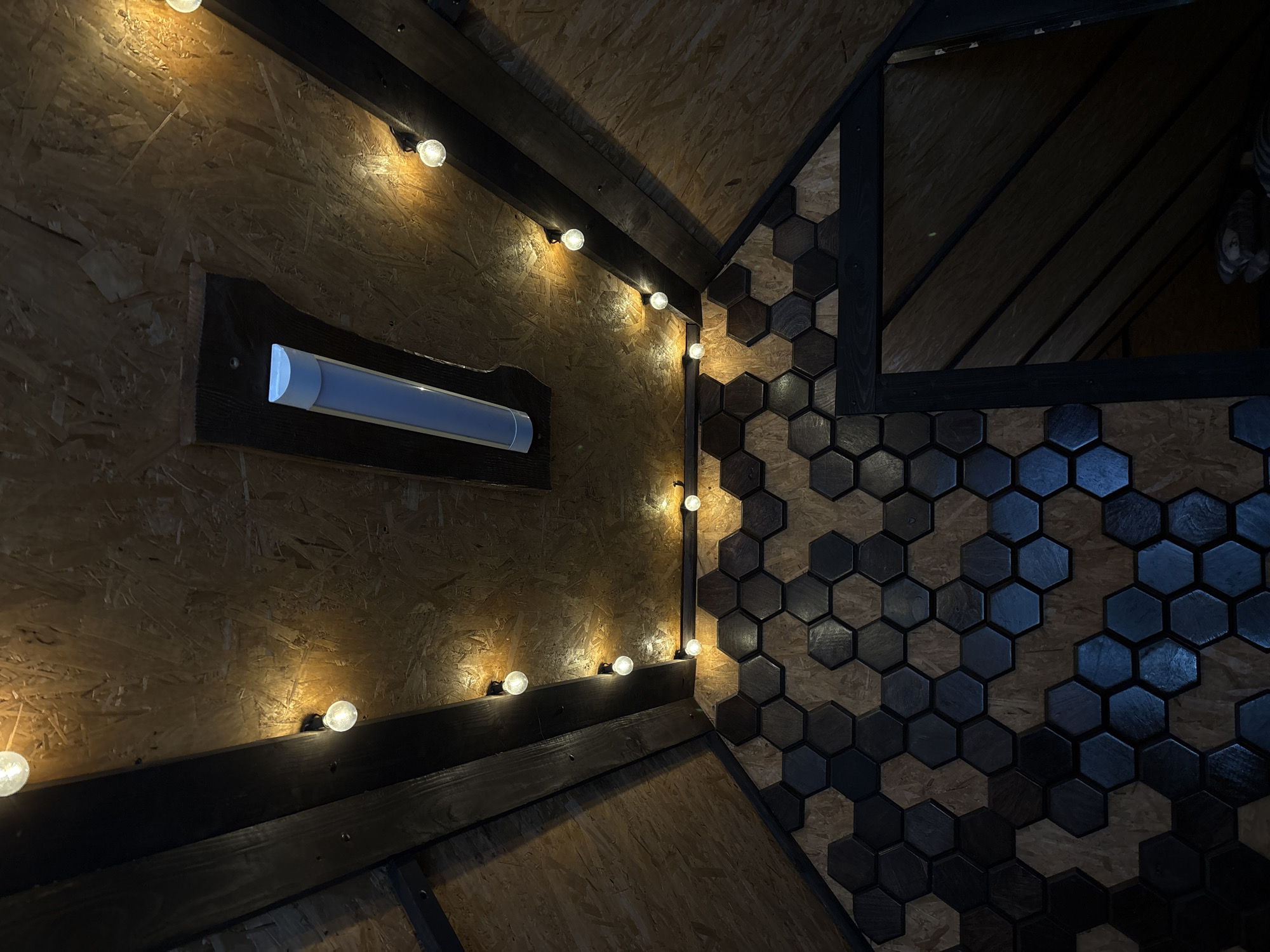 The image shows an interior with a combination of plywood and dark wood hexagon tiles. A string of globe lights runs along the plywood wall, near a rectangular fixture with a fluorescent tube. Dark wood framing is visible. The location is likely Прикамский, Russia, although there is no information in the image to directly confirm this.