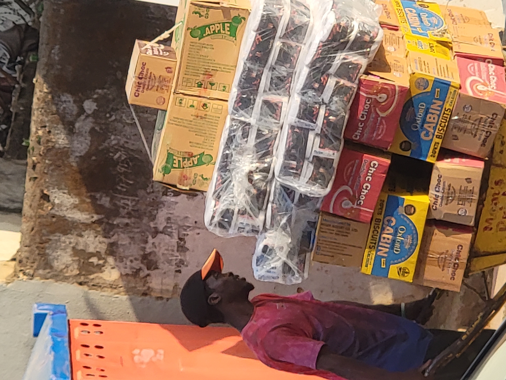 A man in a red shirt and black cap lies on an orange surface. Adjacent to him is a stack of cardboard boxes bearing product names like "APPLE", "Chic Choc Double Cream", and "Oxford CABIN BISCUITS". The location is likely Maiburiji, Nigeria. The surrounding environment shows signs of weathering and possible outdoor storage.