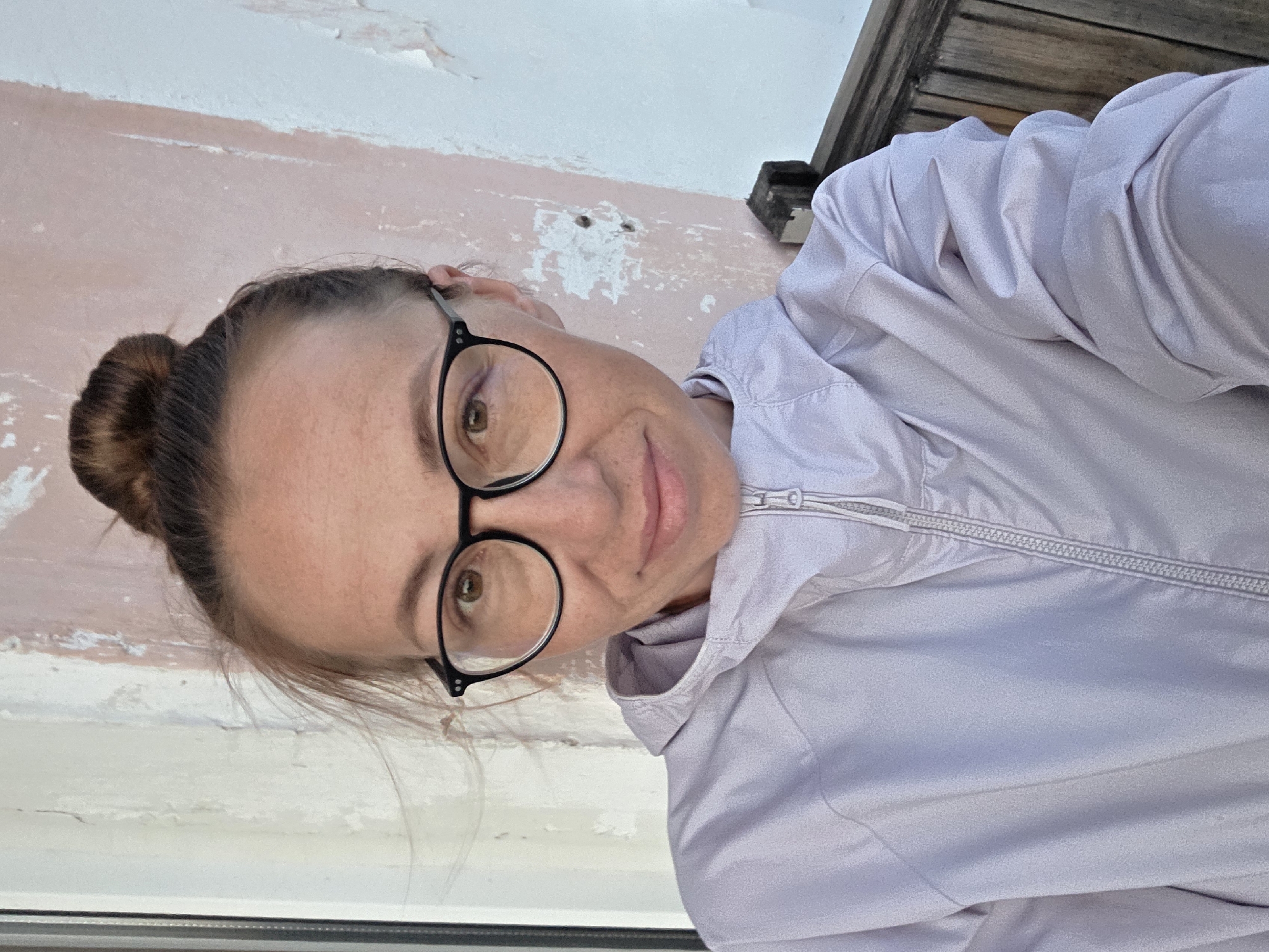 A woman with her hair in a bun and wearing glasses is taking a selfie outdoors. She is wearing a light purple hooded jacket. Behind her is a light-colored wall with peeling paint and a wooden bench. The scene is consistent with an outdoor location in Chaikovsky, Russia, possibly near a residential building or public space.