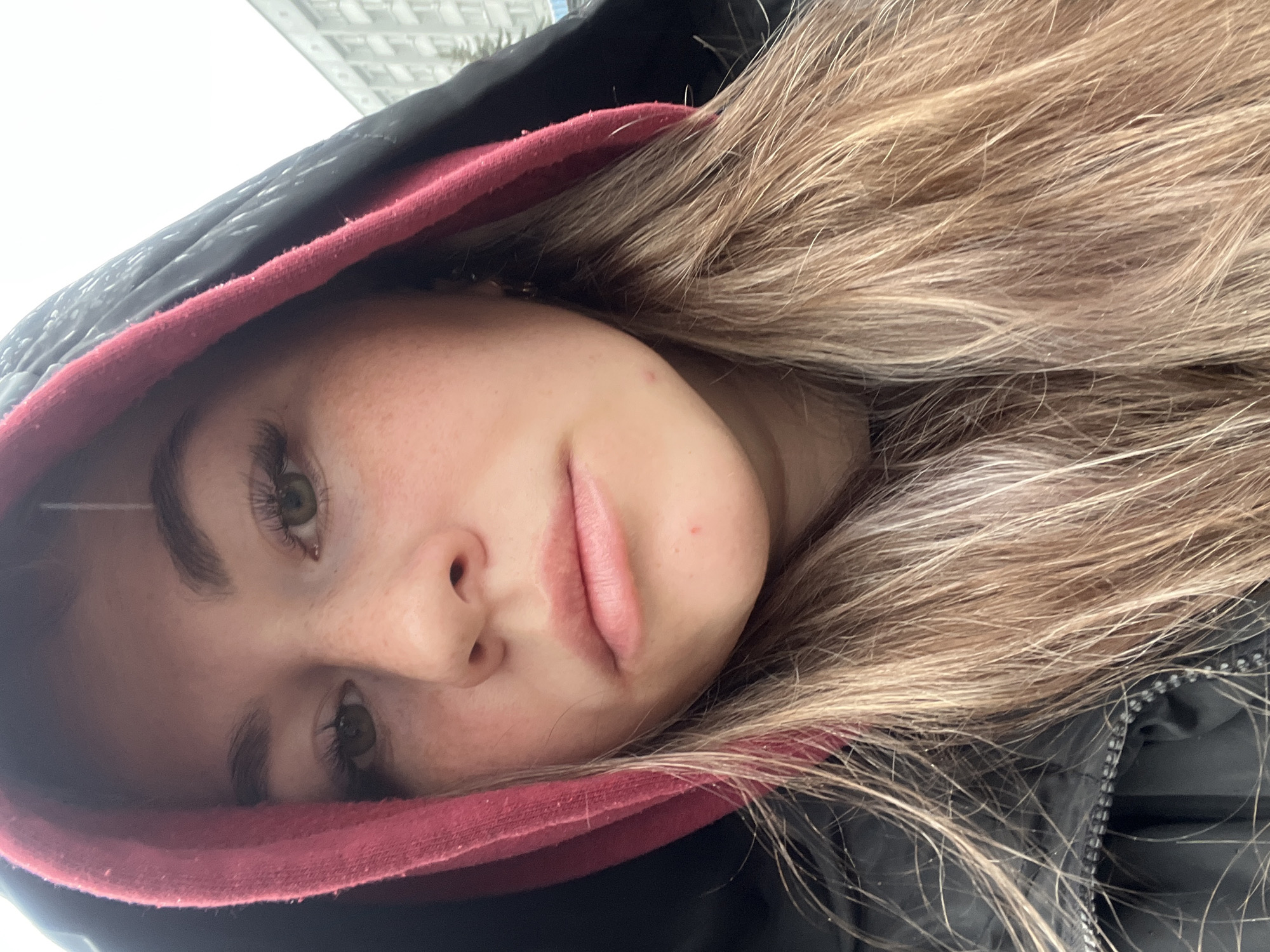 The image is a close-up, diagonal self-portrait of a young woman, captured outdoors. Her face, primarily her left side, occupies a significant portion of the frame, tilted with her gaze directed towards the camera. She has long, light brown or blonde hair that spills out from under a black hooded jacket, which features a prominent red inner lining.
Her expression is natural and relaxed, with a subtle curve to her full lips suggesting a slight, soft smile. Her eyes appear to be a light color, possibly hazel or green, and her eyelashes are prominent, hinting at mascara or extensions. Her skin shows a natural texture with some visible freckles or minor blemishes.
The background suggests an overcast daytime setting, with diffused light illuminating her face. A bright, light gray sky fills the upper background, against which a distant, indistinct building or structure can be faintly seen. The hooded jacket implies a cool or chilly environment. No other individuals or specific objects are visible, and there is no discernible text within the image. The provided context indicates the location as Chaikovsky, Russia.